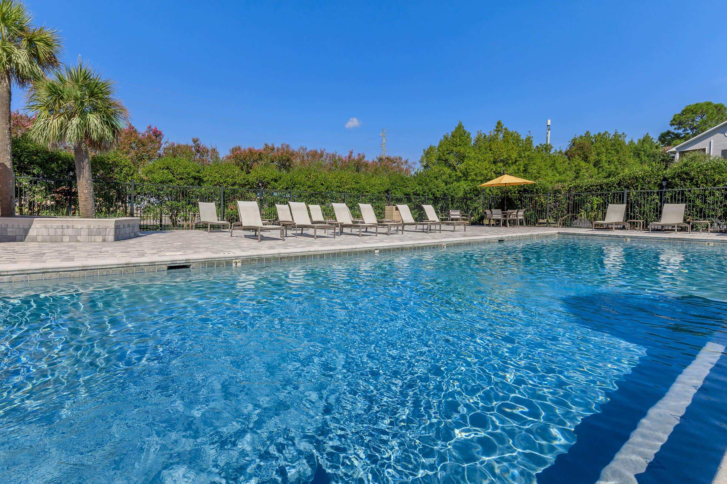 A clear swimming pool surrounded by lounge chairs on a sunny day. Lush greenery and trees are visible in the background, along with a sunshade umbrella. The sky is bright blue with a few fluffy clouds, creating a relaxing outdoor atmosphere.