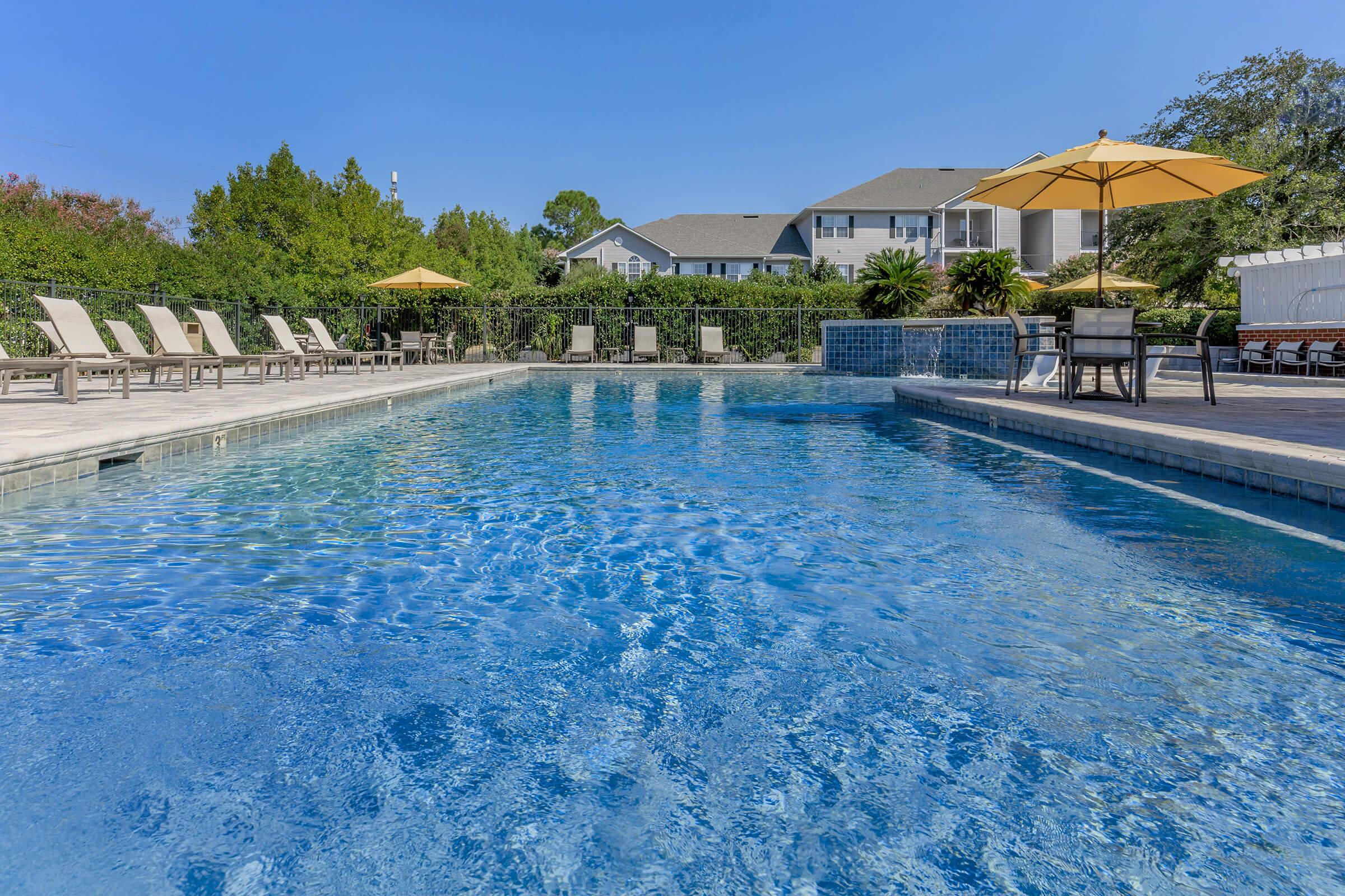 A sunny pool area featuring clear blue water, surrounded by lounge chairs and yellow umbrellas. In the background, there are lush green trees and a residential building, creating a relaxing outdoor atmosphere perfect for leisure and enjoyment.