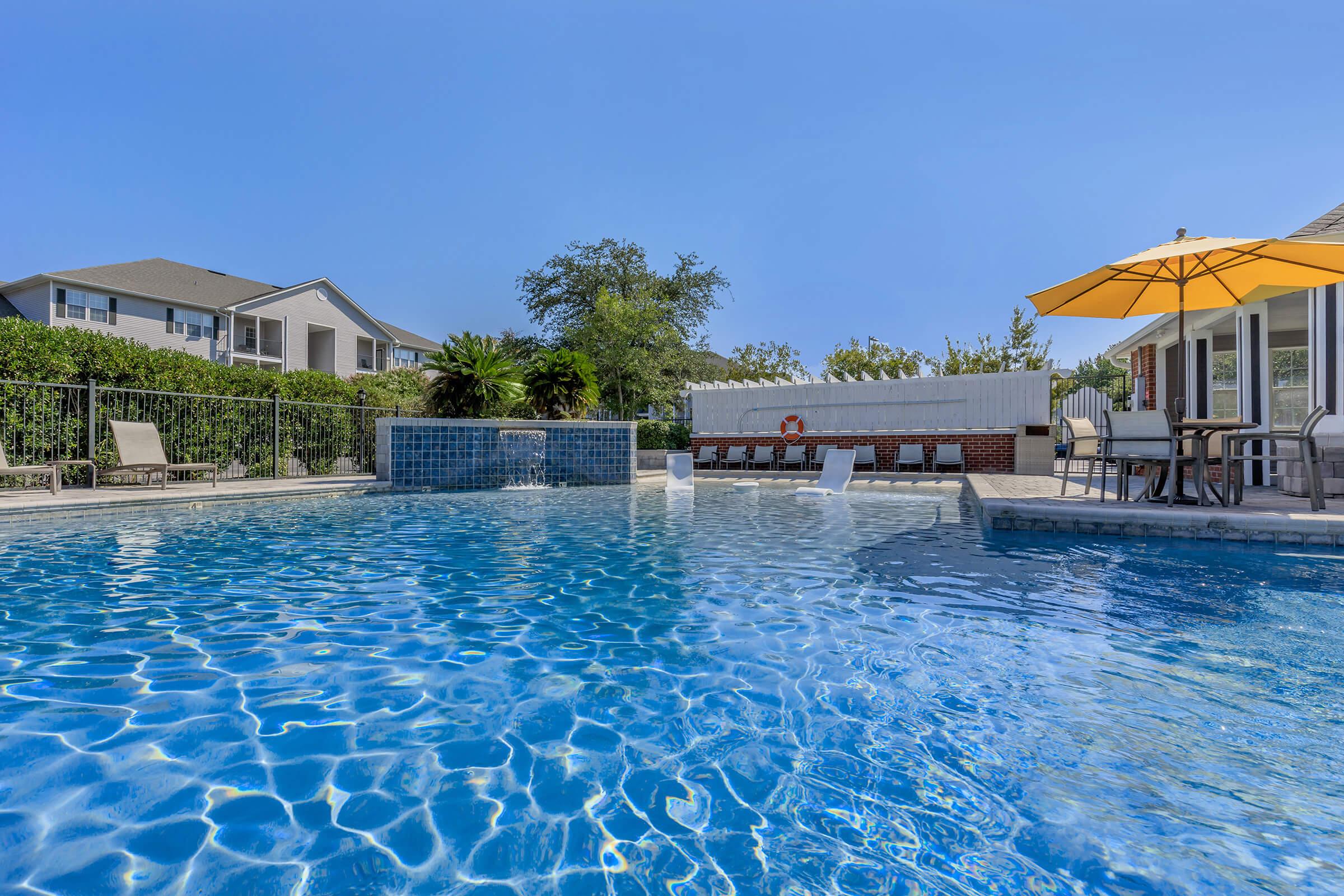 A clear blue swimming pool surrounded by lounge chairs and a large yellow umbrella. In the background, residential buildings and lush greenery are visible. The scene is bright and sunny, evoking a relaxing atmosphere perfect for leisure and enjoyment.