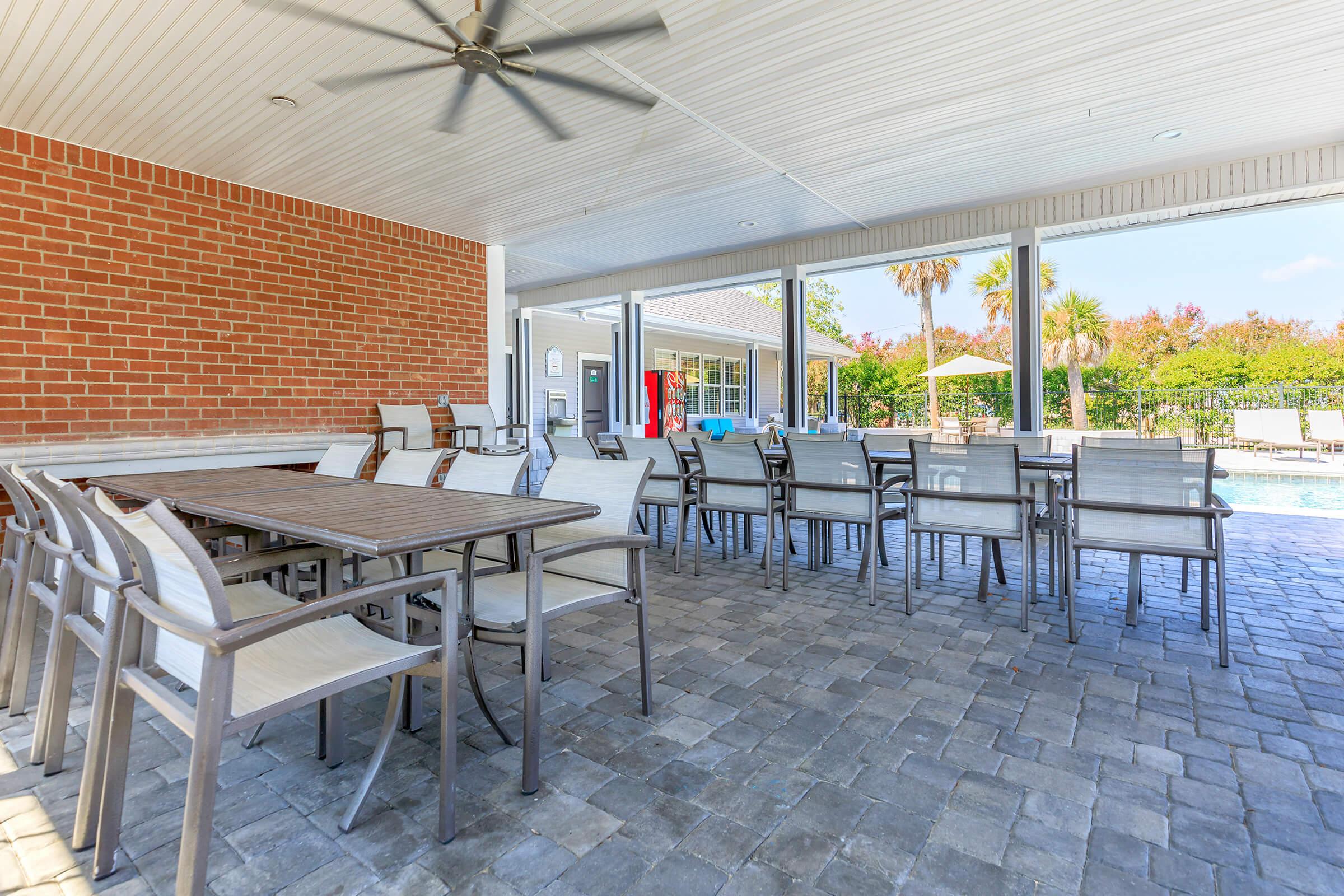 An outdoor seating area with multiple tables and chairs under a covered patio. Features a brick wall and a ceiling fan, with a view of a swimming pool and palm trees in the background. The space is designed for relaxation and social gatherings.