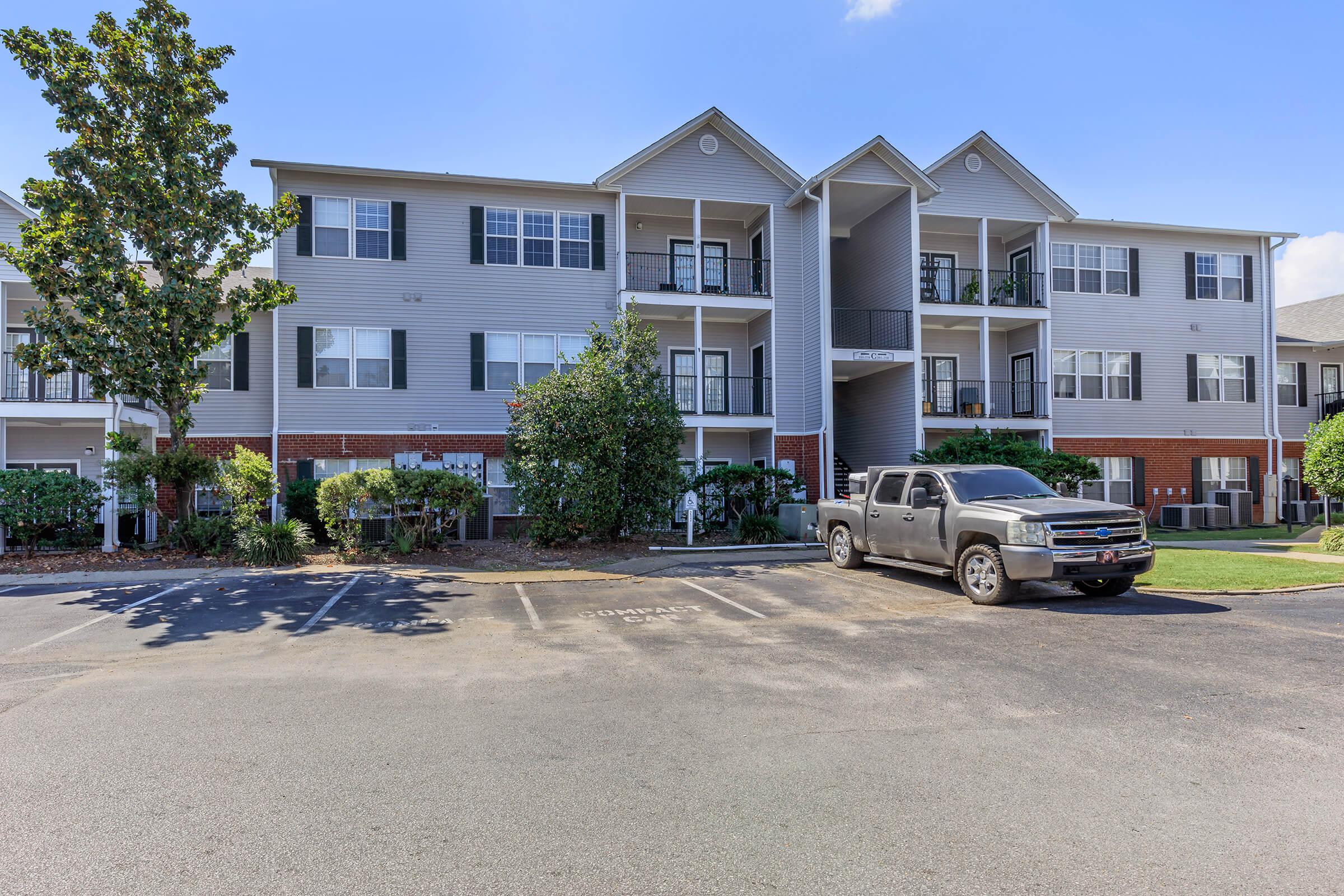 A multi-unit residential building with a beige facade and green shutters. The structure features multiple balconies and has landscaping with shrubs. In the foreground, a silver pickup truck is parked on a paved lot. There are designated parking spaces visible. Blue sky and sunlight enhance the scene.