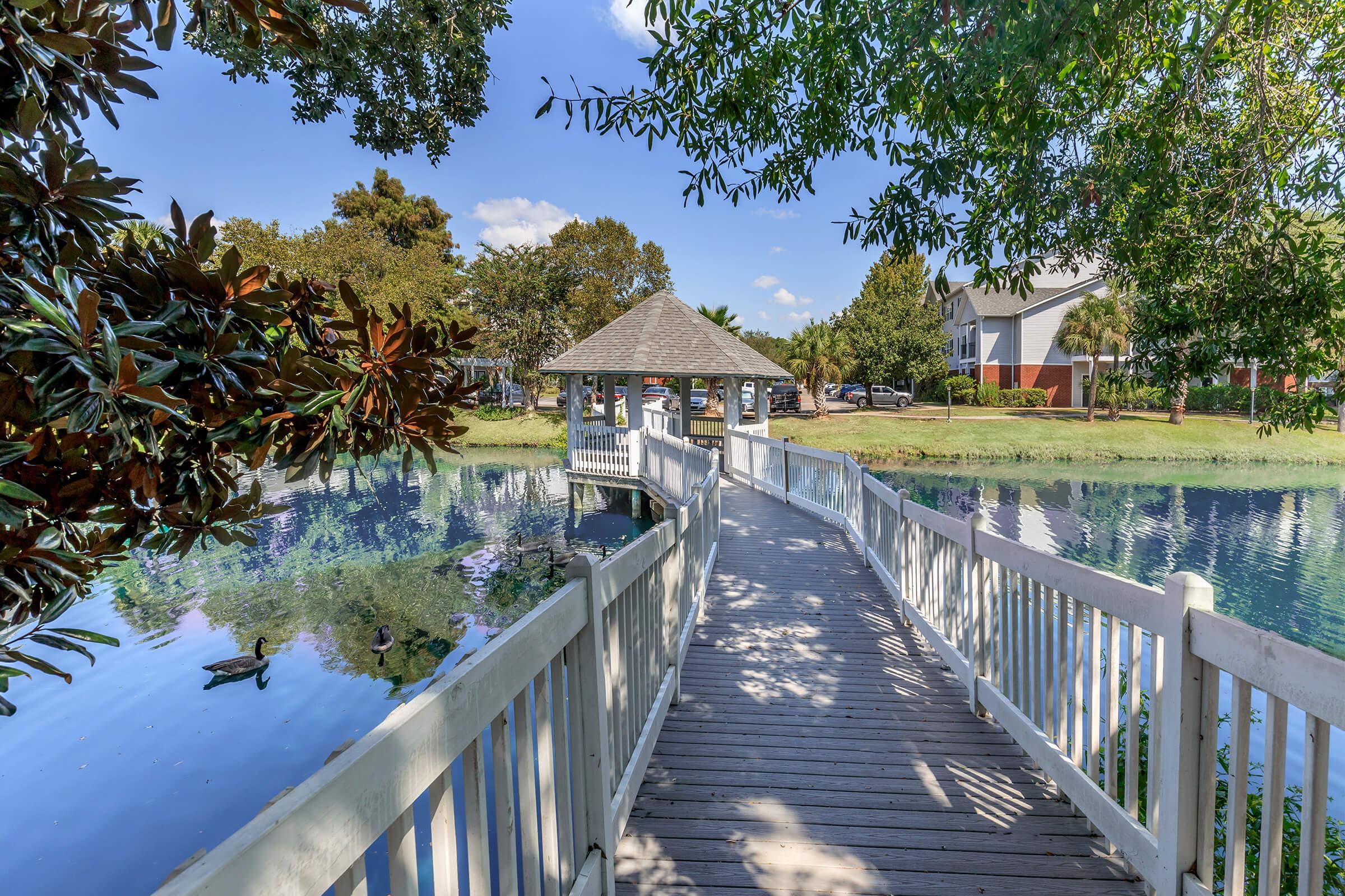 A wooden walkway leads to a gazebo overlooking a serene pond, surrounded by lush green trees. The water reflects the blue sky and fluffy clouds, creating a peaceful outdoor scene.
