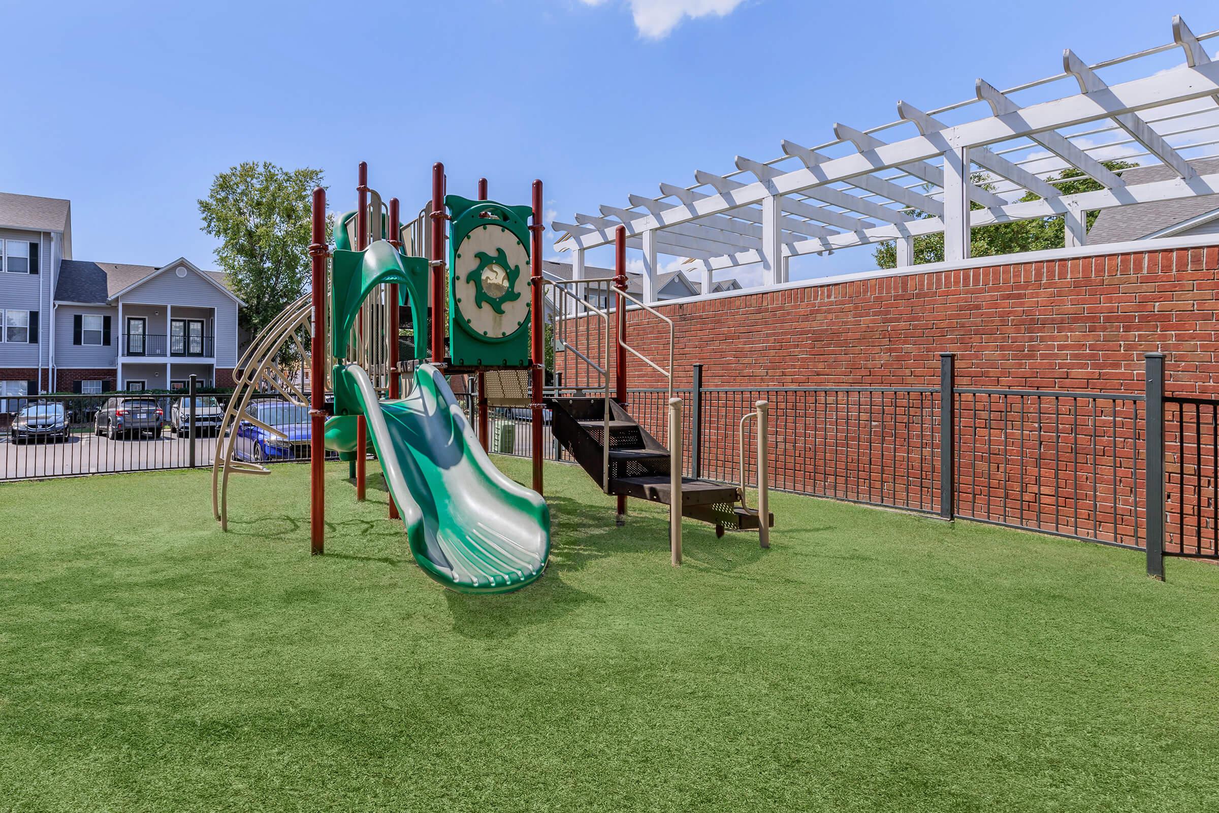 A playground featuring a green slide and climbing structure surrounded by grass. The background includes a brick wall and a covered area with a trellis, along with several parked cars visible in the distance. The setting is sunny and inviting for children to play.