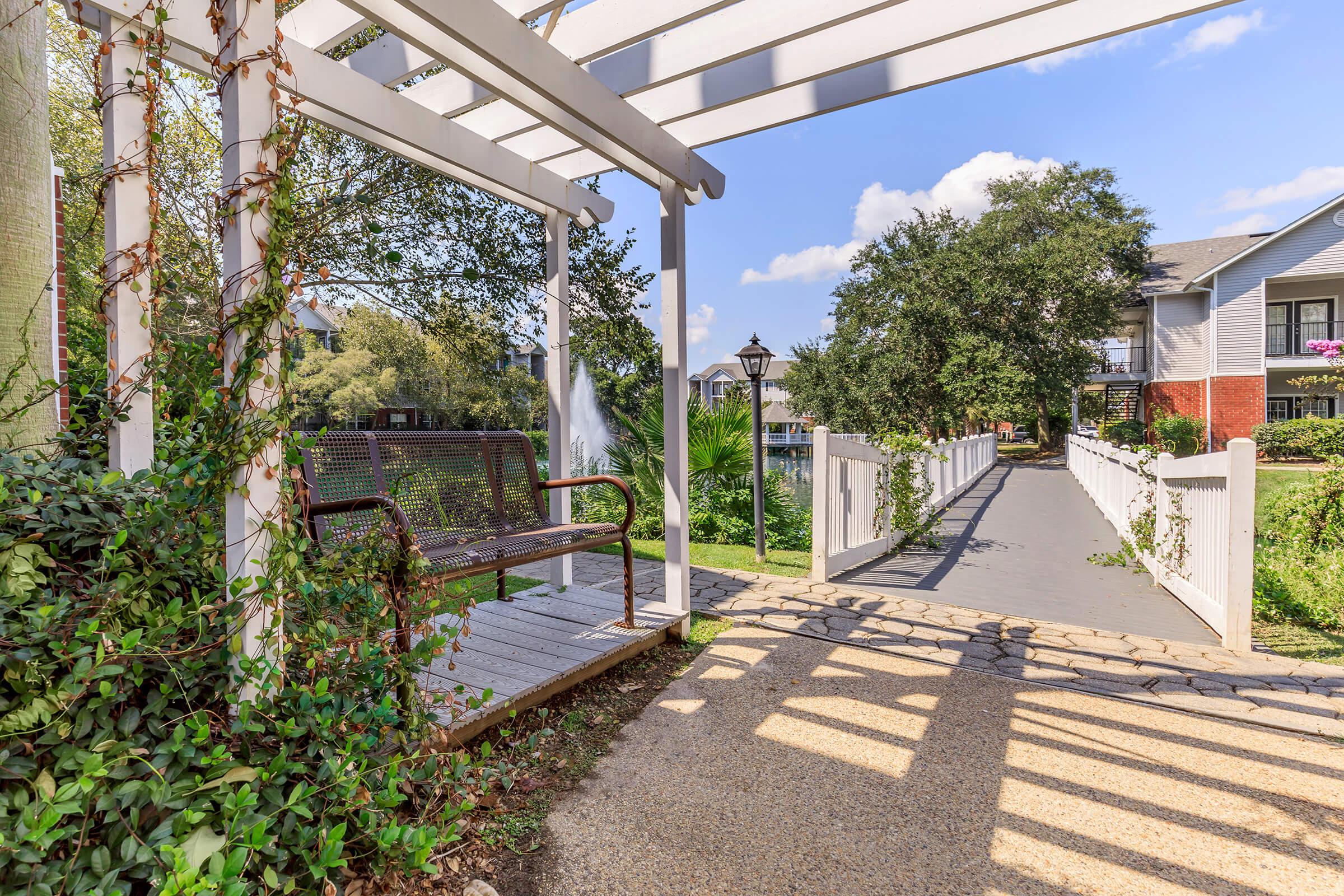 A peaceful pathway lined with greenery, featuring a wooden bench under a white pergola. The scene includes a lamp post and a well-maintained walkway leading to nearby buildings, with blue skies and fluffy clouds overhead, creating a serene outdoor environment.