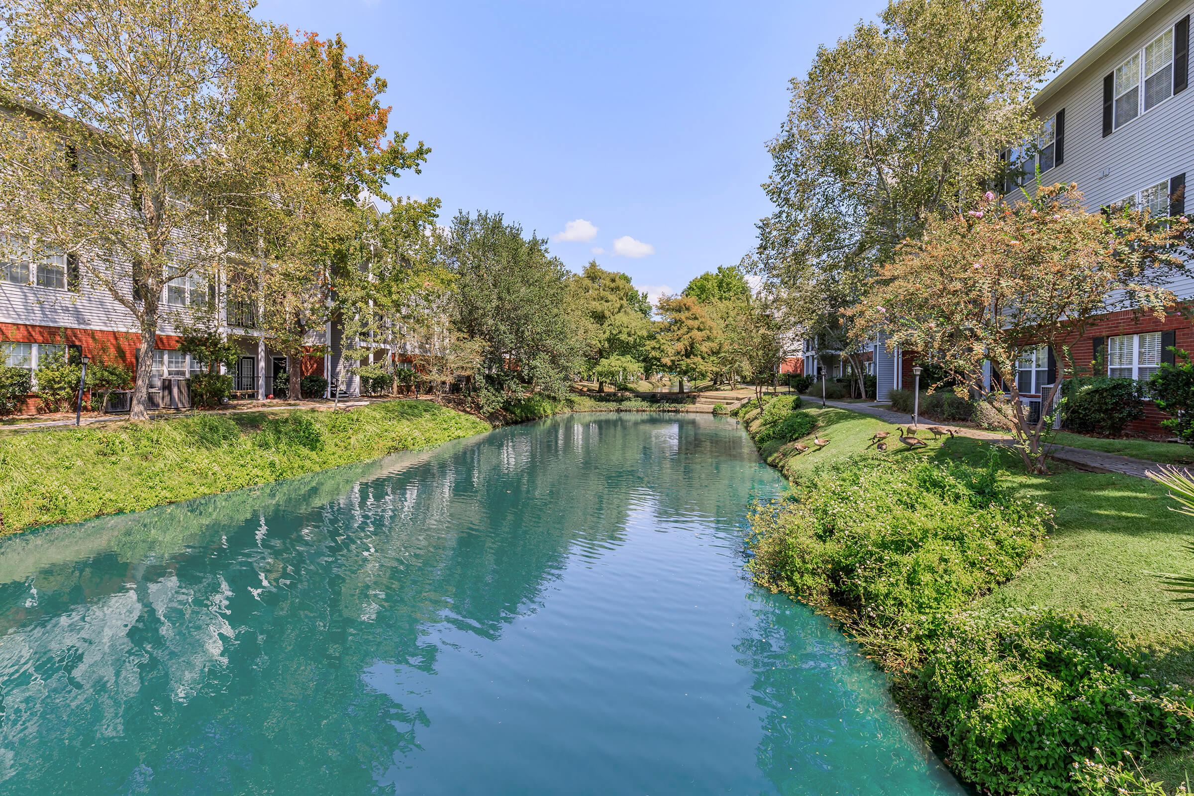 Serene view of a narrow, turquoise waterway surrounded by lush greenery and trees, with landscaped banks. On one side, there are residential buildings with white and red accents, creating a peaceful and inviting atmosphere. The clear sky adds to the tranquil setting.