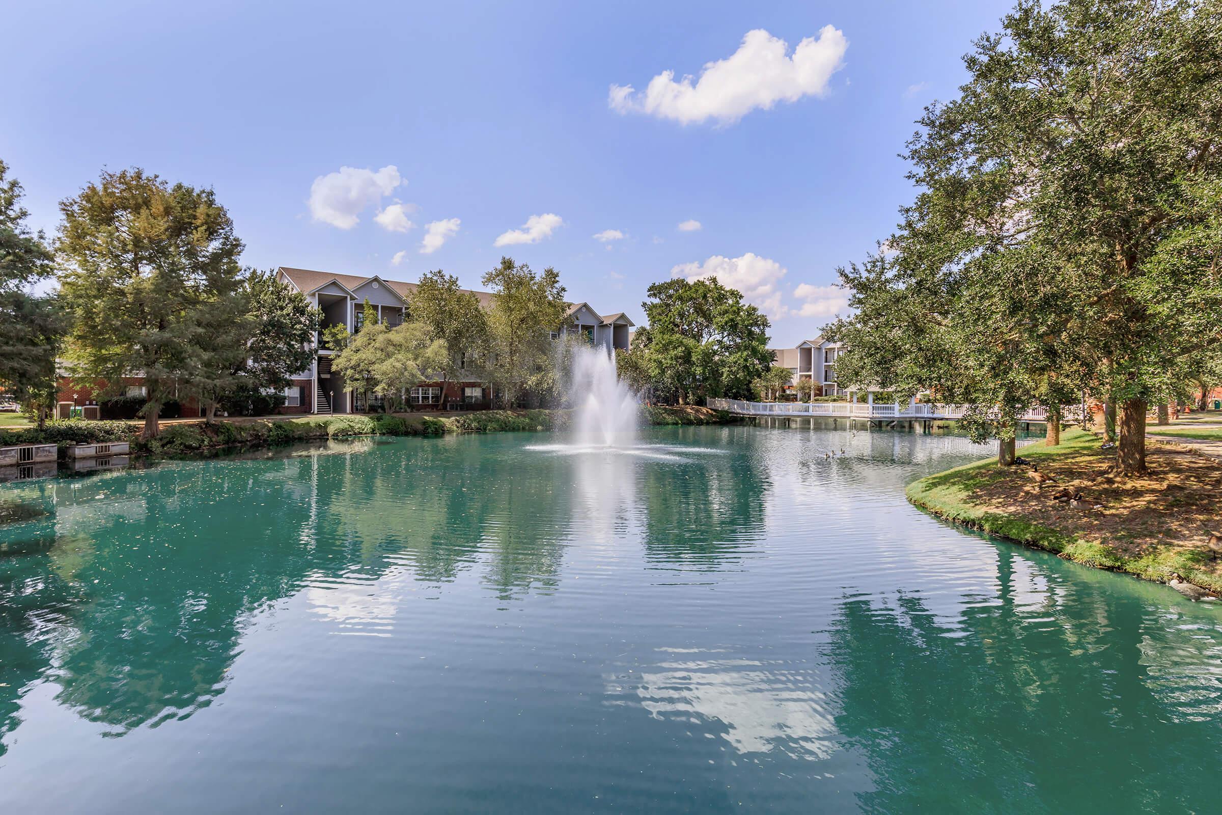 A serene pond surrounded by lush trees and residential buildings. A fountain sprays water into the air, creating ripples on the calm surface. Reflections of the blue sky and clouds enhance the tranquil scene, inviting a sense of peace and relaxation.