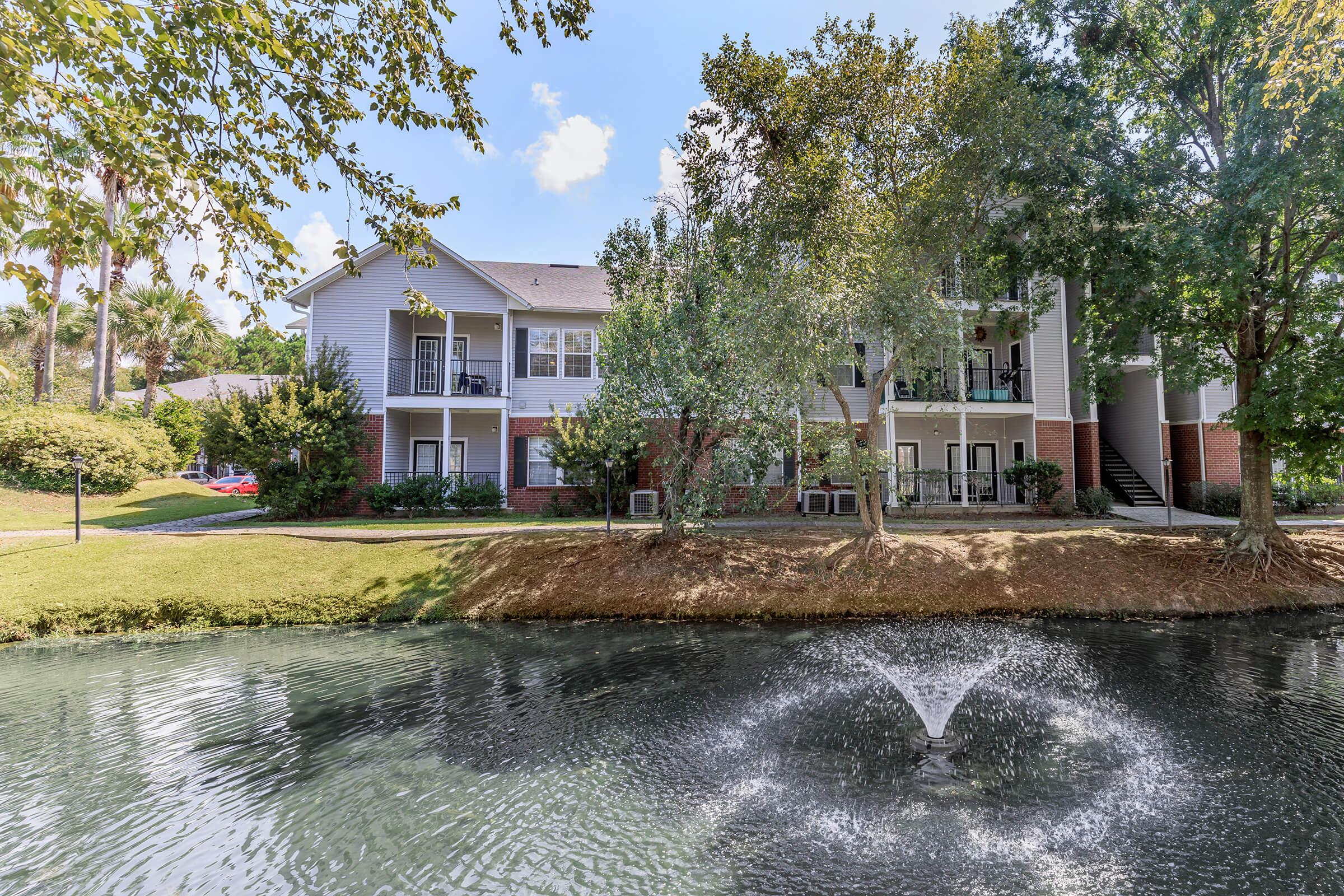 A multi-story apartment building surrounded by greenery, with a small pond in the foreground. A fountain creates a gentle water spray in the pond, adding to the serene atmosphere. The building features balconies, and the scene is bright and inviting under a clear sky.