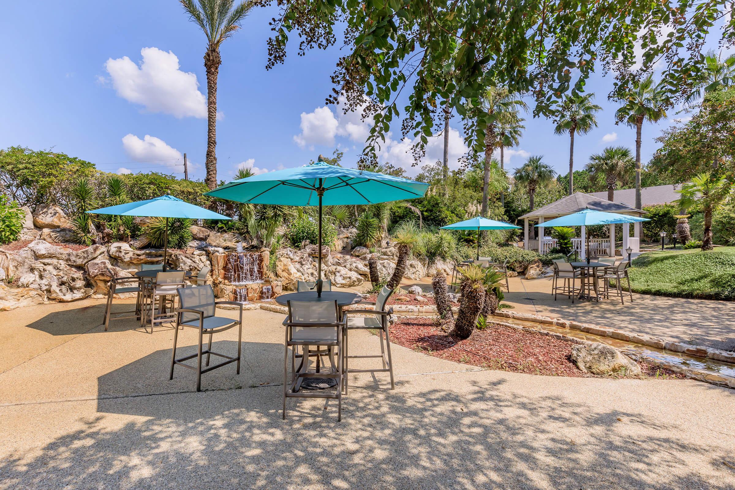 A sunny outdoor lounge area featuring several tables with turquoise umbrellas, surrounded by lush greenery, palm trees, and a rock waterfall. The pathway is sandy, and there is a gazebo in the background, creating a relaxing atmosphere for guests.