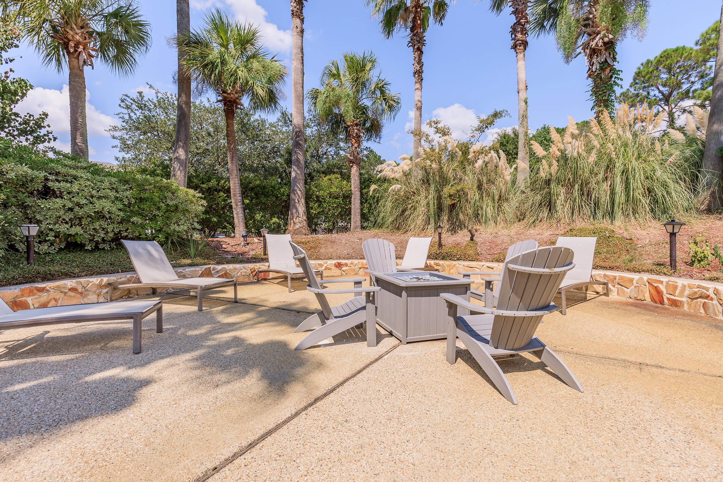 A serene outdoor patio featuring several gray Adirondack chairs and a small table, surrounded by lush green palm trees and ornamental grasses under a clear blue sky. Sunlight casts gentle shadows on the textured surface, creating a relaxing atmosphere perfect for leisure and gatherings.