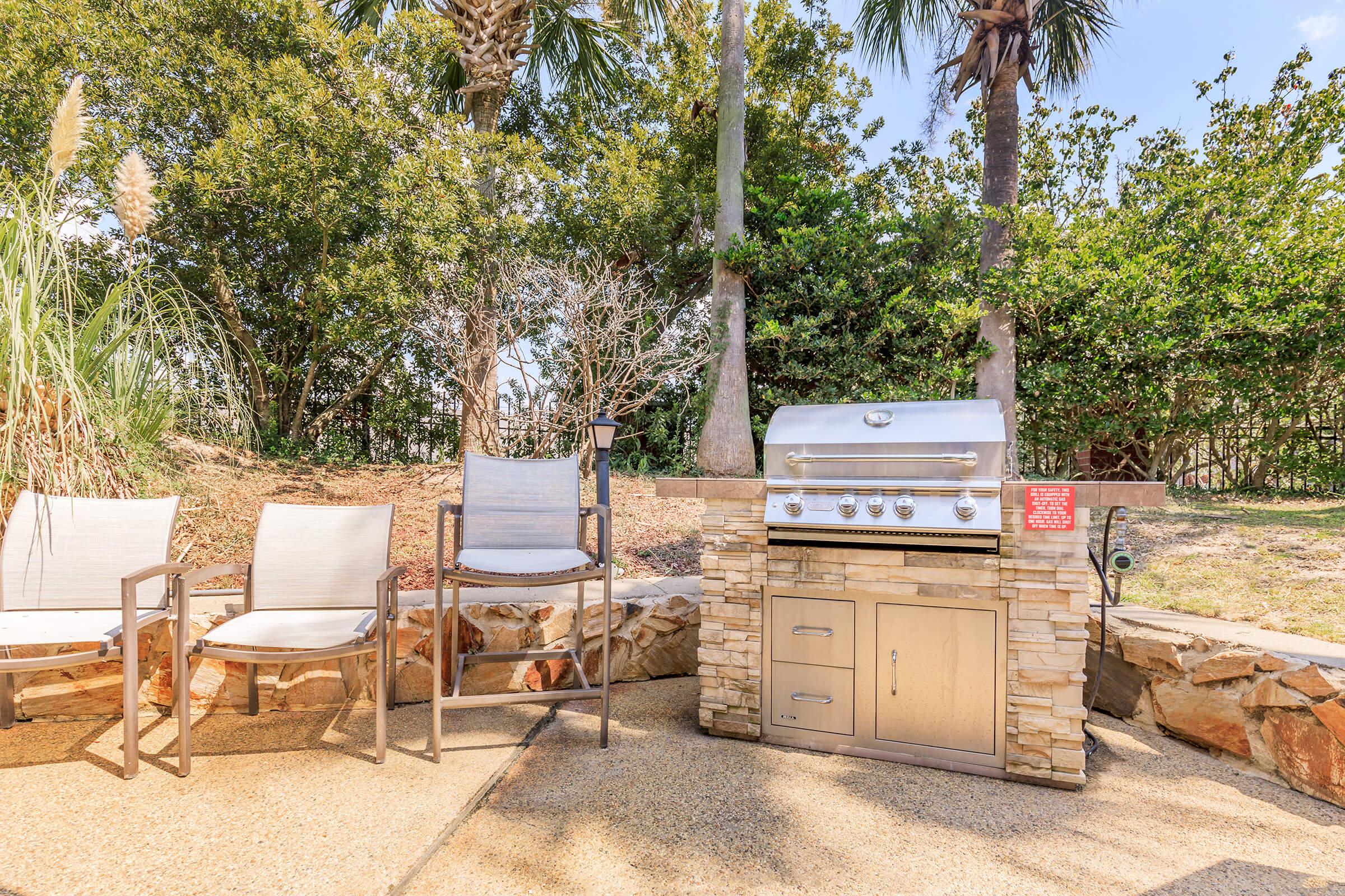 A modern outdoor barbecue grill situated beside two metal chairs on a stone patio. The grill is silver with multiple burners and a storage compartment below. Surrounding greenery includes palm trees and other plants, creating a relaxed outdoor atmosphere.
