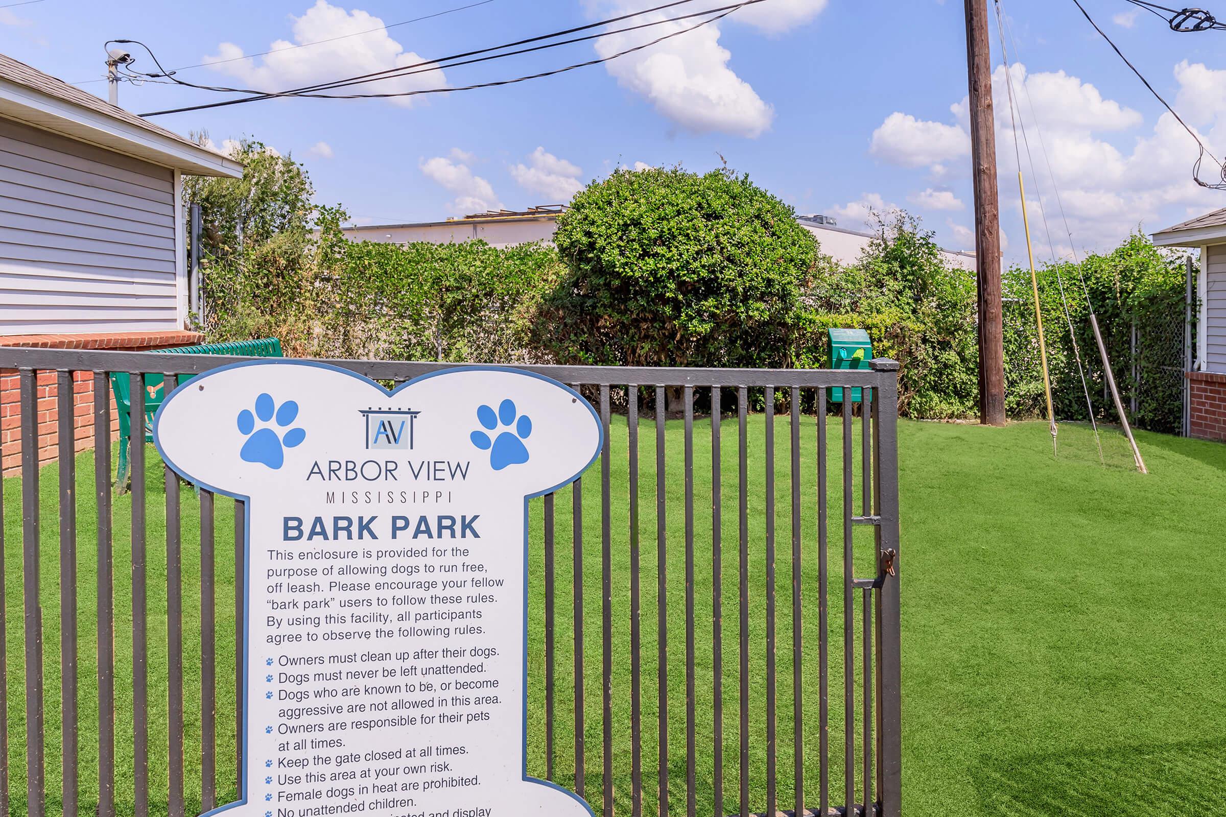 A fenced area labeled "Bark Park" at Arbor View, Mississippi, with a sign outlining rules for dog owners. The park features artificial grass, nearby trees, and swings in the background. The scene is set under a clear blue sky with fluffy clouds.