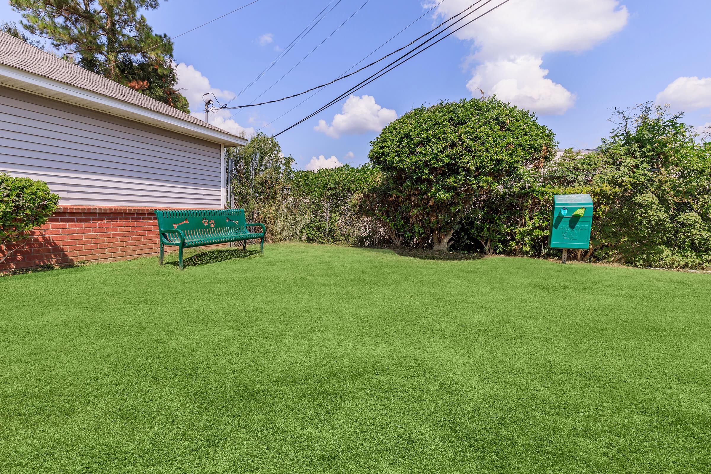 A grassy area featuring a green bench near the side of a house. The landscape includes a bushy hedge and a mailbox in the background. Blue sky with fluffy clouds is visible overhead, creating a serene outdoor setting.