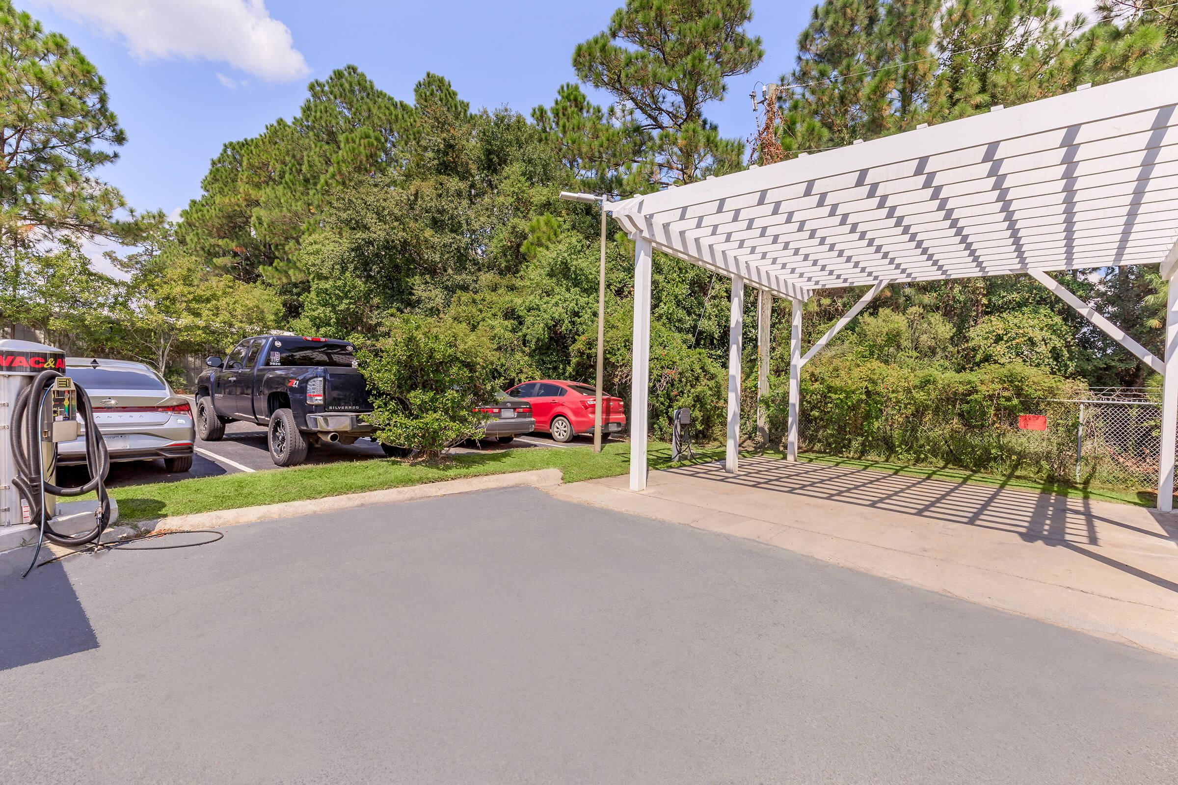 Parking area showcasing two vehicles, one red and one black, under a white pergola. Surrounding greenery and trees provide shade, with a clear blue sky in the background. The scene is well-lit, indicating a sunny day.