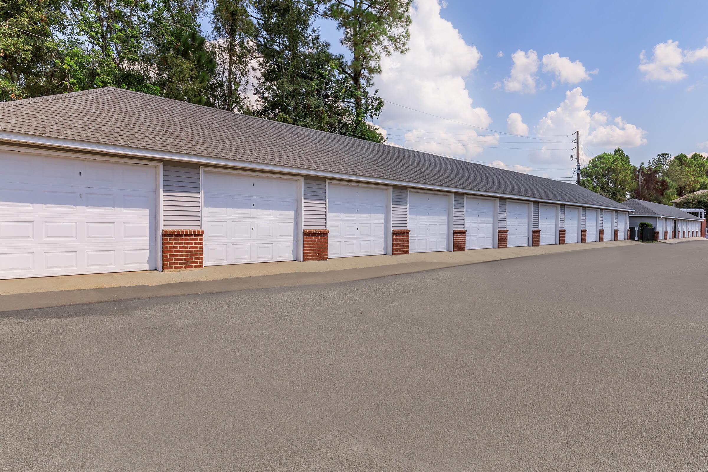 A row of storage units with white garage doors, set against a clear blue sky with a few clouds. The units are lined up neatly along a paved road, surrounded by greenery in the background.