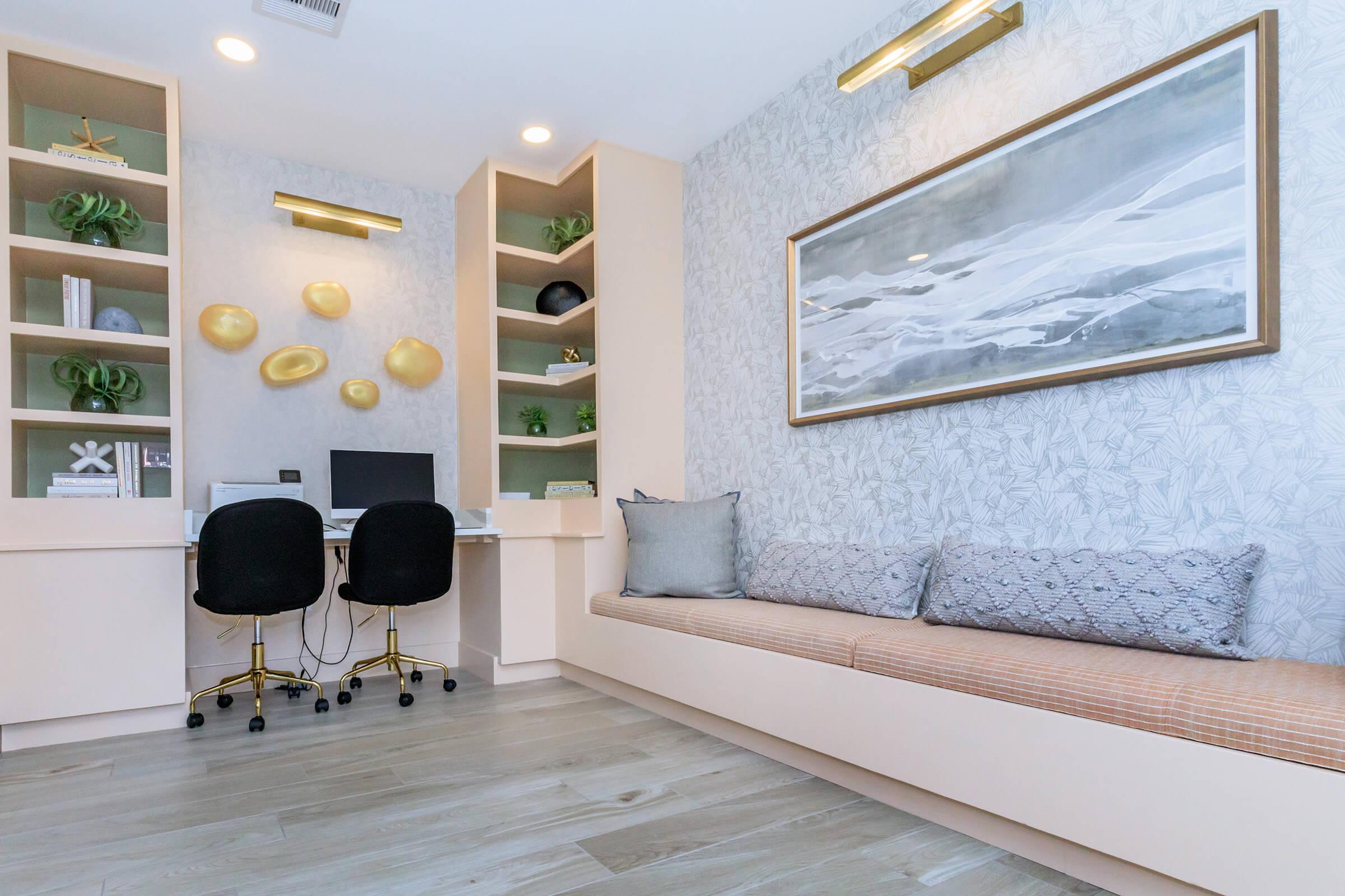 A modern home office featuring a minimalist design. It includes a built-in desk with a computer, two black chairs, and shelves showcasing decorative items. A cozy bench with patterned cushions is set against a light-colored wall adorned with abstract art and stylish gold accents.