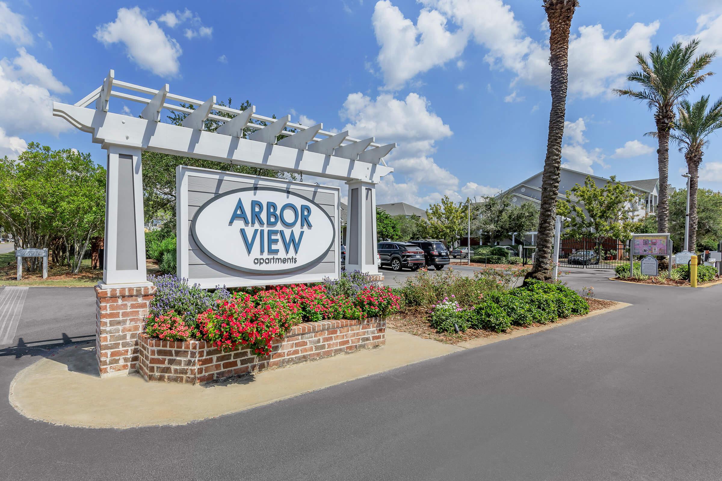 Signage for Arbor View Apartments, featuring a large oval sign with the text "ARBOR VIEW" in a prominent position. The area is landscaped with colorful flowers and palm trees, and there are parked cars in the background. The sky is bright with fluffy clouds, creating a welcoming atmosphere.