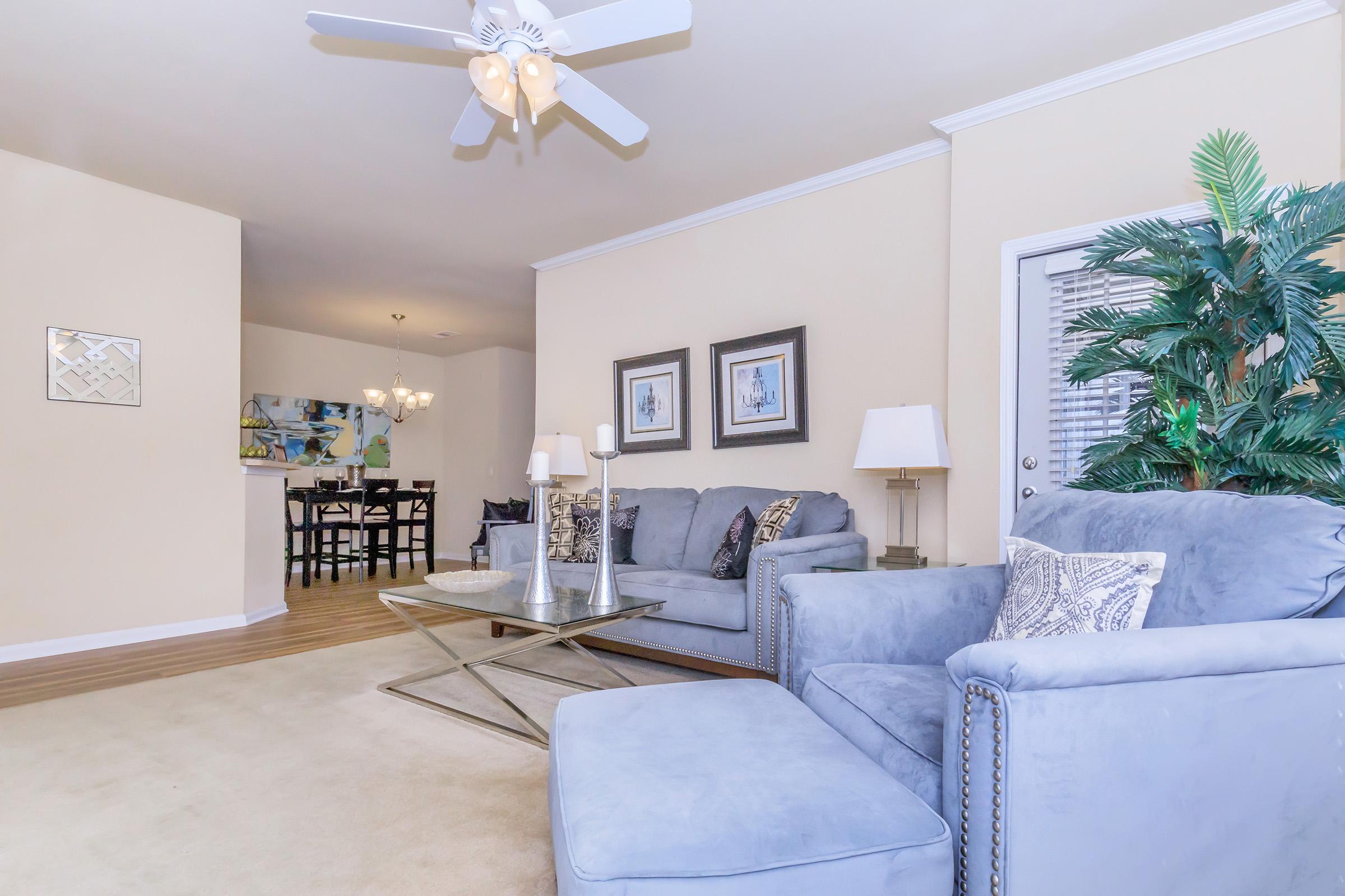 A cozy living room featuring a light blue couch and armchair with decorative pillows, a coffee table in the center, and two lamps on side tables. A potted plant adds a touch of greenery, and a dining area is visible in the background with a chandelier and a modern painting on the wall.