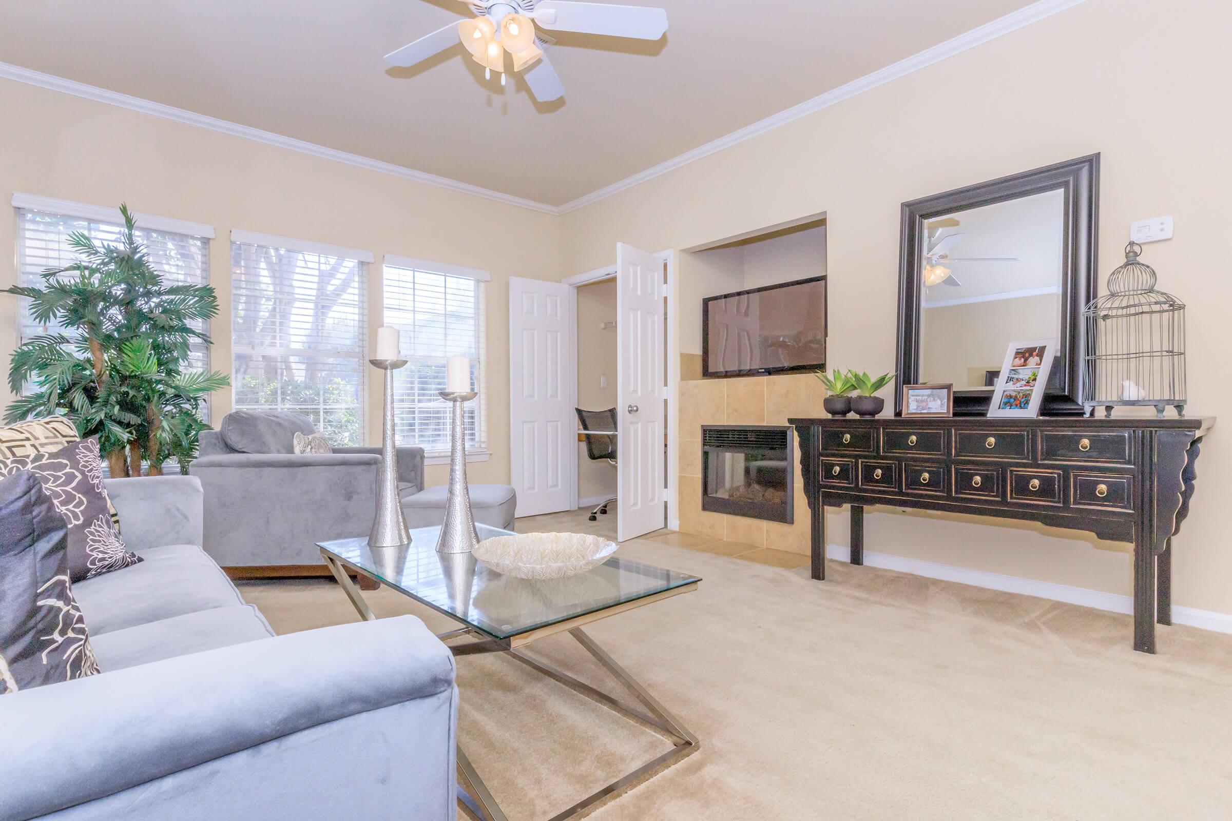 A cozy living room featuring a gray sofa, a glass coffee table, and decorative plants. There’s a stylish wooden console table against the wall, with candles and a decorative birdcage. Sunlight filters through the windows, creating a warm and inviting atmosphere. A fireplace is visible in the background.