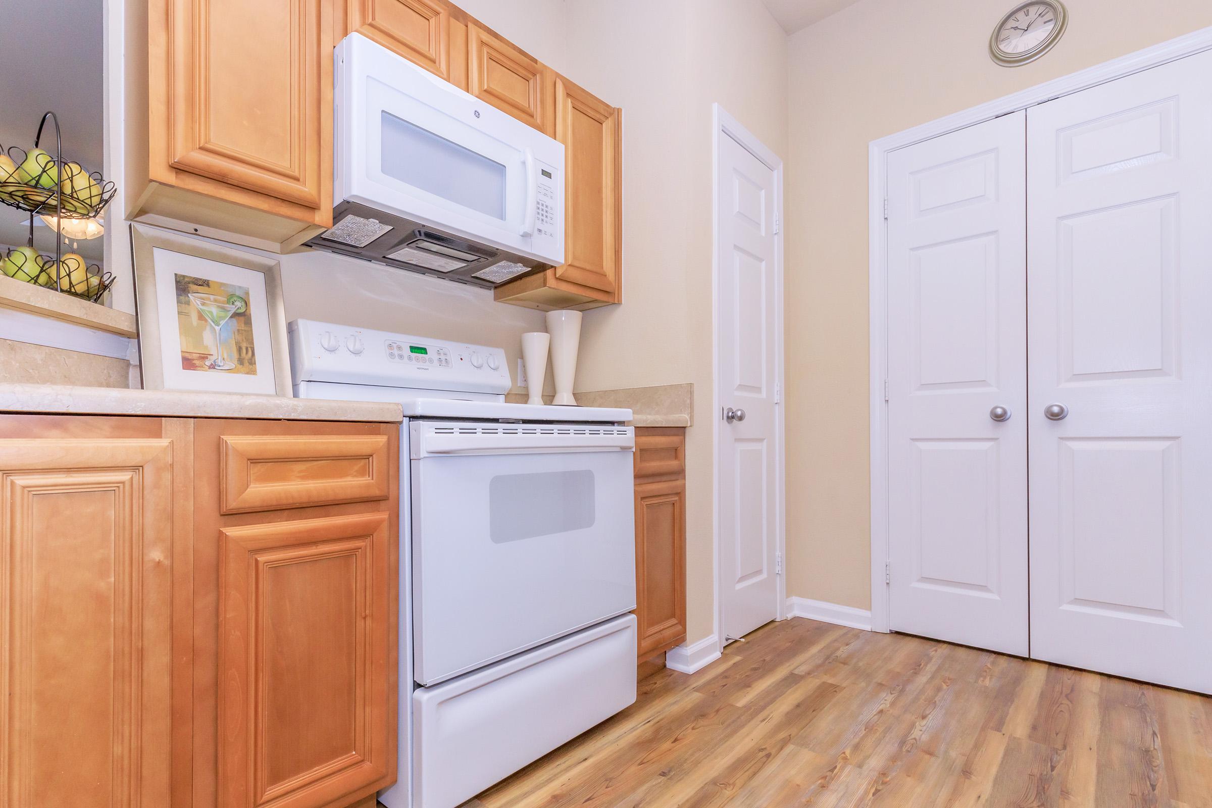 A modern kitchen featuring light wooden cabinets, a white stove, and a microwave above it. The countertop is neat with a framed picture and two vases. A clock is mounted on the wall, and a closed closet is visible on the right with double doors. The floor has a warm wood-like finish.