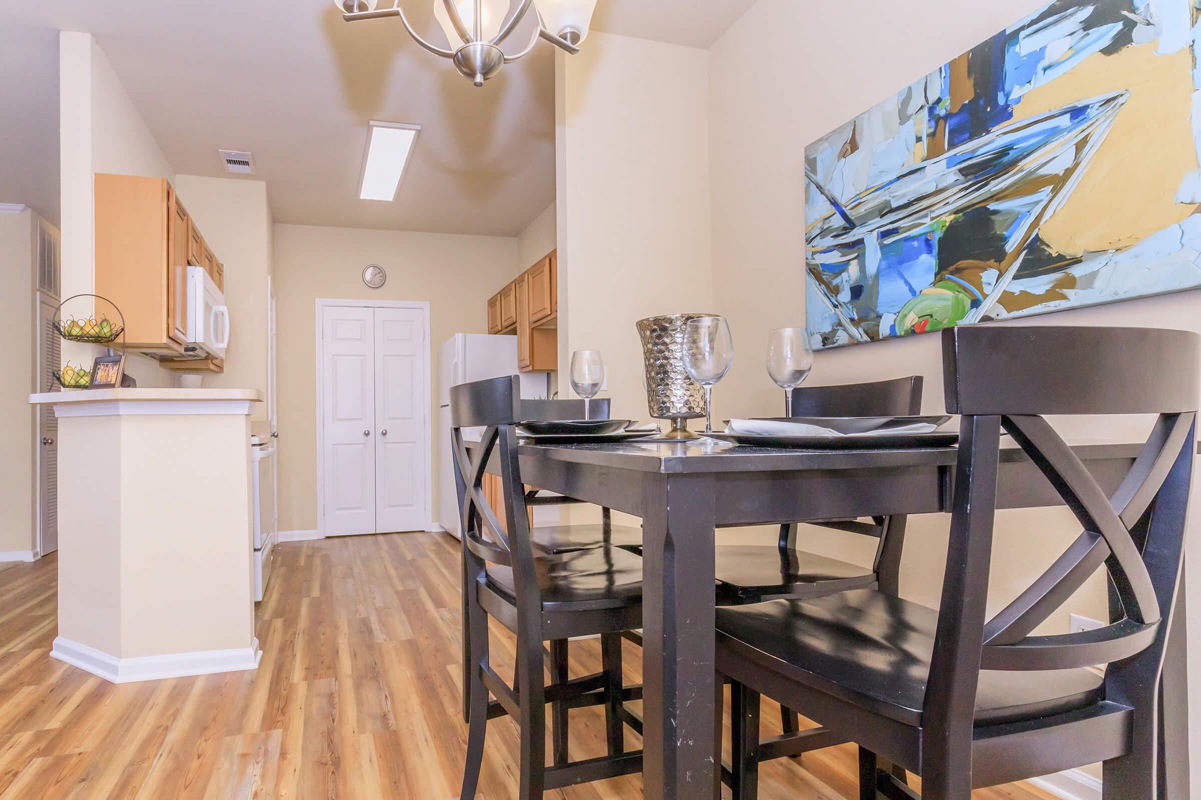 A dining area featuring a black table and chairs set for two with wine glasses. A modern abstract painting hangs on the wall above the table. The kitchen area is visible in the background, showcasing wooden cabinetry and appliances. Warm wooden flooring completes the cozy atmosphere.