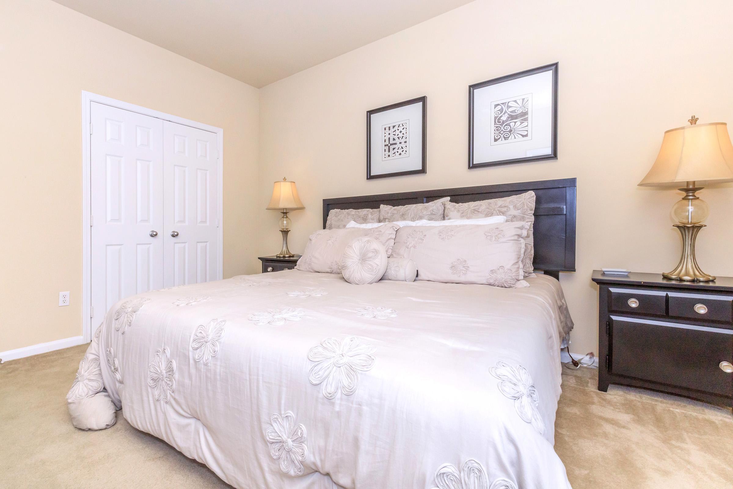 A cozy bedroom featuring a large bed with decorative pillows and a light-colored quilt, flanked by two bedside tables with lamps. The walls are painted a soft beige, and two framed artworks are hung above the bed. A white closet door is visible in the background, and the floor is covered in light carpet.