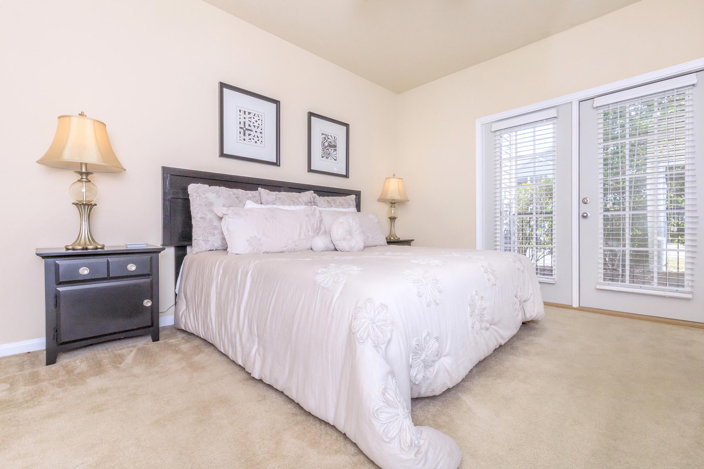 A softly decorated bedroom featuring a large bed with a light-colored quilt and decorative pillows. Two bedside lamps provide soft lighting, and framed artwork adorns the walls. A pair of double doors with blinds opens to a view outside, and the room has beige walls and light carpet.