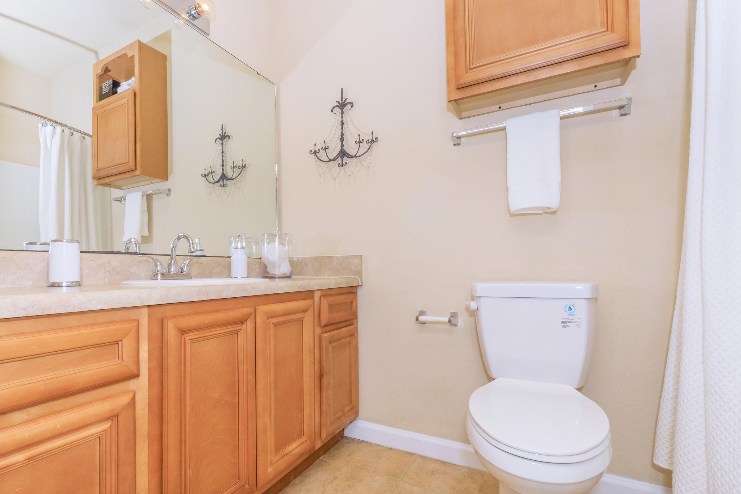 A clean bathroom featuring a beige wall, wooden cabinetry, a white toilet, a sink with a countertop, and a large mirror. There are decorative wall elements and neatly arranged towels, creating a tidy and inviting space.