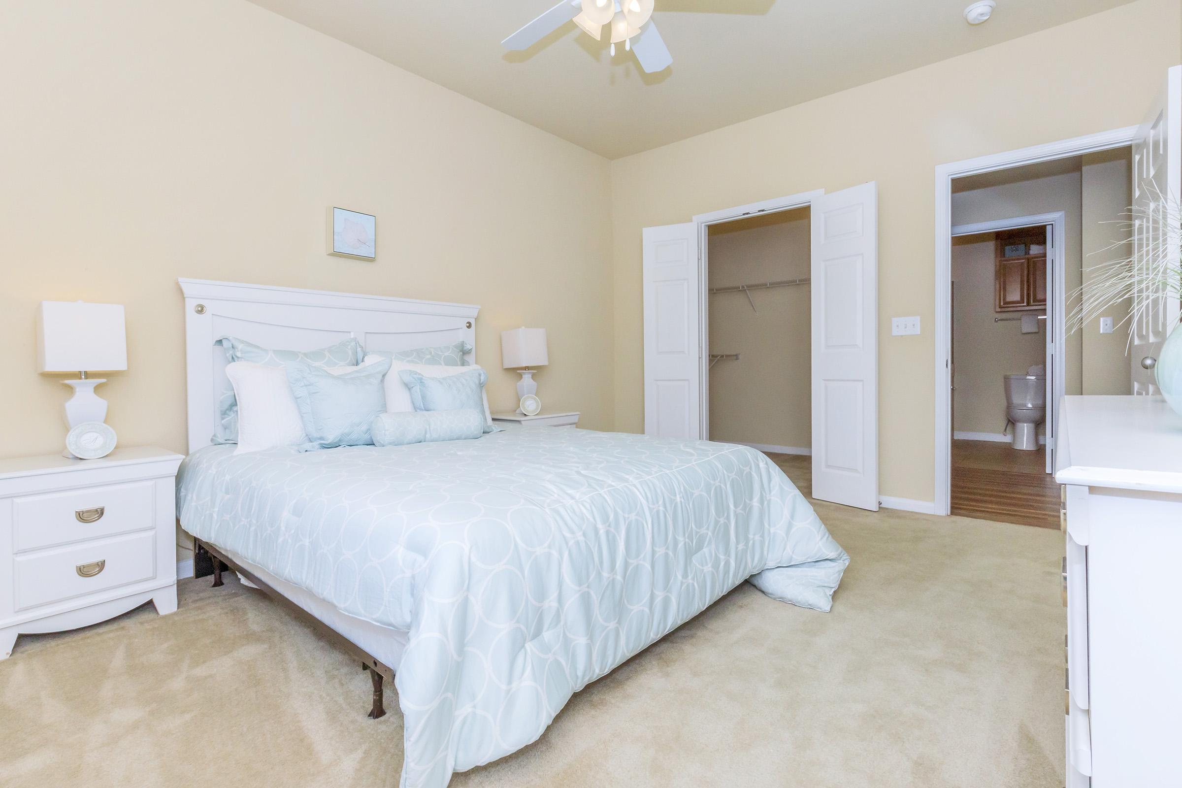 A cozy bedroom featuring a light blue comforter on a queen-sized bed with several decorative pillows. Two white bedside tables with lamps are on either side of the bed. The walls are painted a soft beige, and there's a closet with double doors in the background. A door leads to another room, and the carpet is beige.