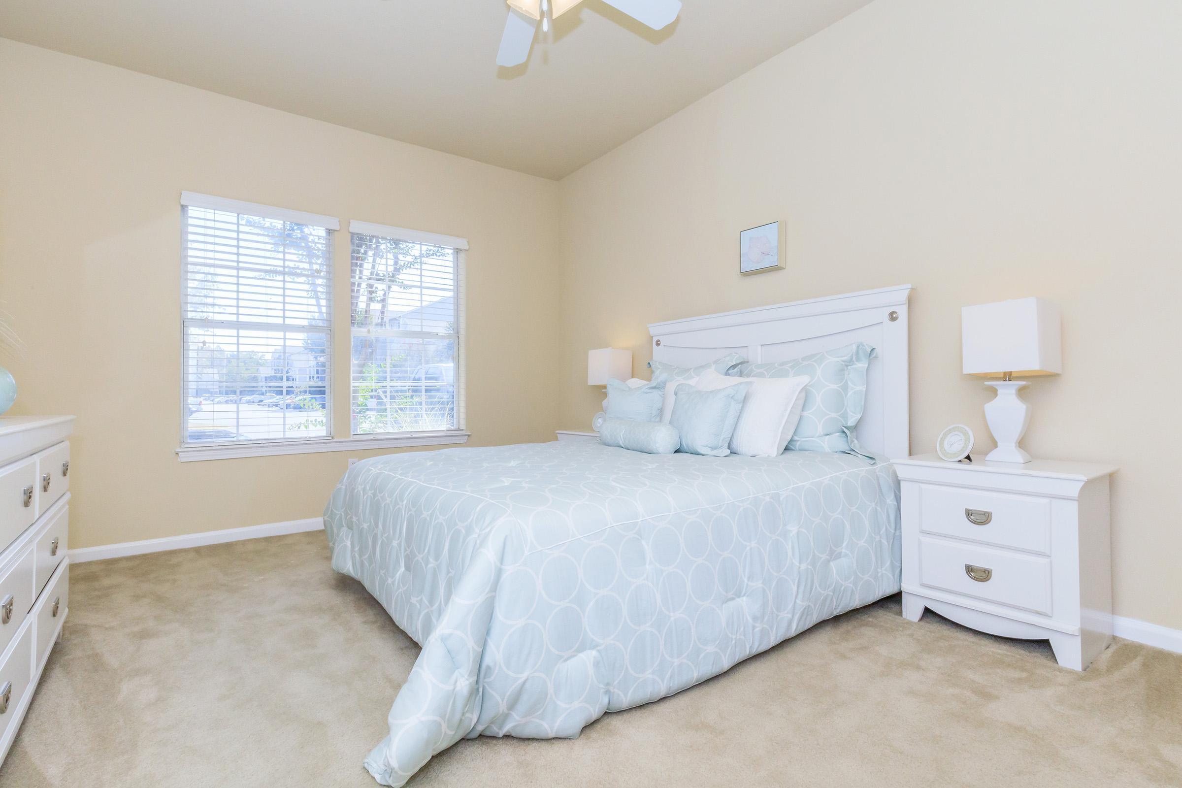 A cozy bedroom featuring a queen-sized bed with light blue bedding and decorative pillows. The walls are a soft beige, and there's a white dresser and bedside table. A window with blinds allows natural light to enter, highlighting the warm, inviting atmosphere. Soft carpet covers the floor.