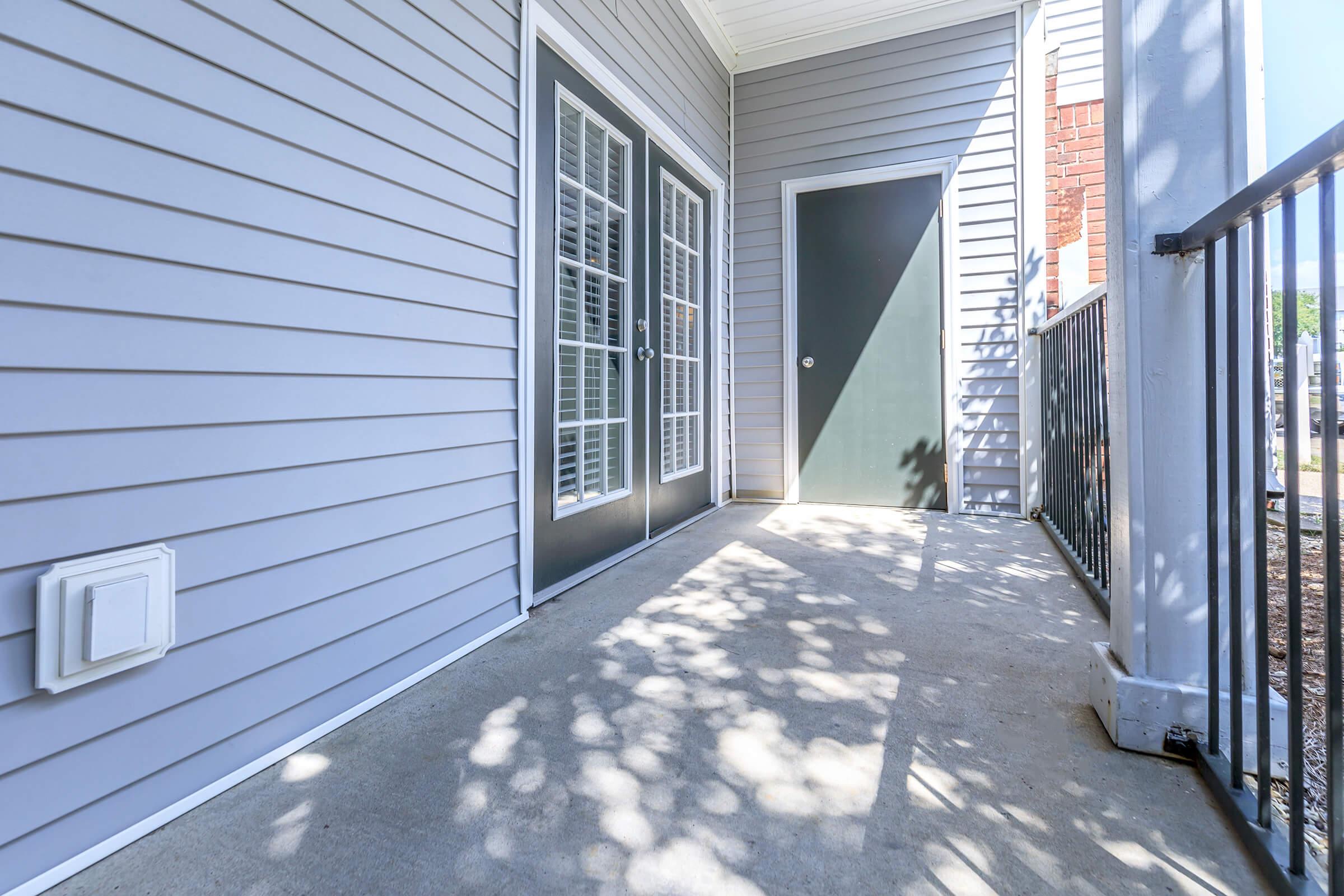 A perspective view of a porch with a concrete floor and gray siding walls. Two double doors with glass panels are visible on the left, while a single green door is on the right. The area is well-lit, with shadows from nearby trees creating patterns on the floor.