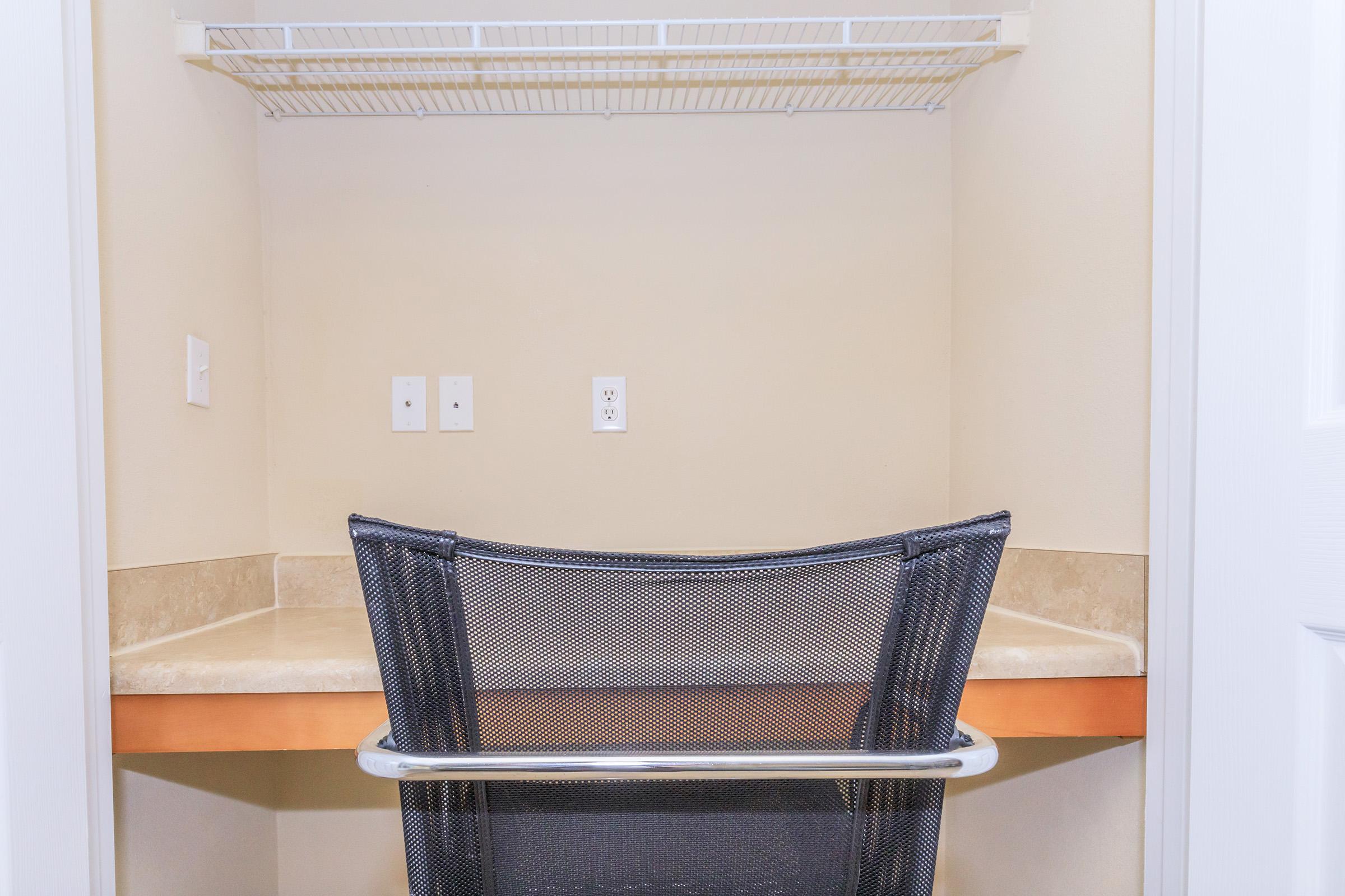 A small, empty workspace with a beige wall. There is a simple countertop and a black mesh chair positioned in front of it. Above the workspace, there is a wire shelf. Two electrical outlets are visible on the wall next to the countertop. The space appears tidy and well-organized.