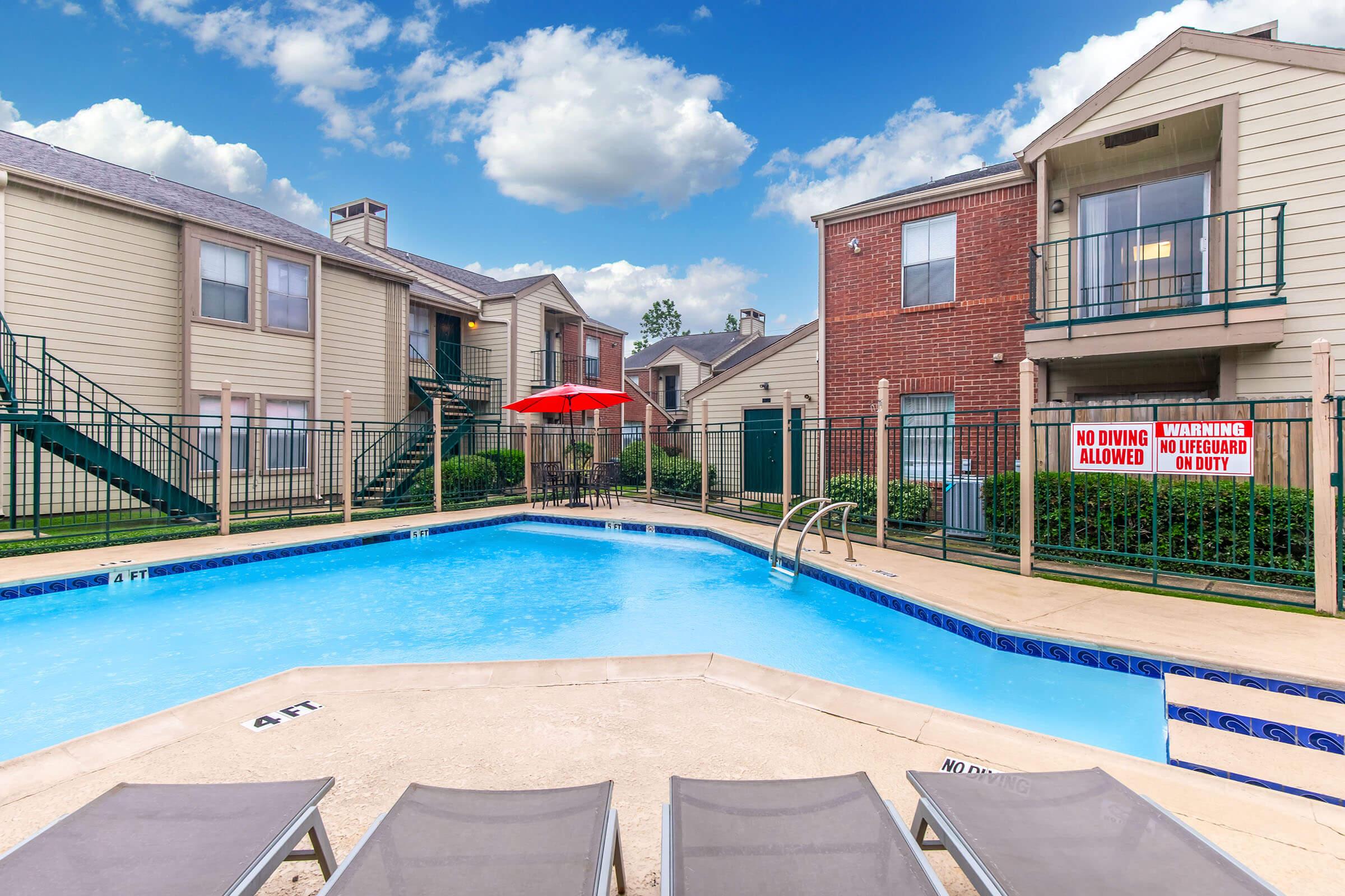 A clean, inviting swimming pool surrounded by lounge chairs. Two multi-story apartment buildings are visible in the background, with green staircases and a red umbrella providing shade. There are warning signs posted near the pool area, and a bright blue sky with fluffy clouds above.
