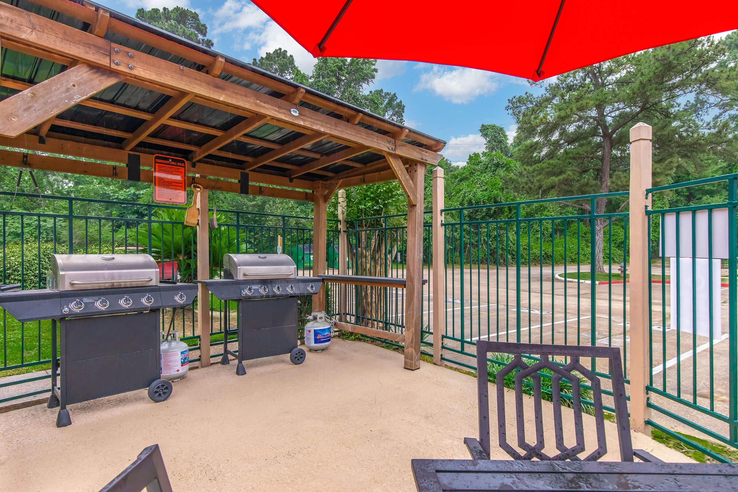 A shaded outdoor grilling area featuring two stainless steel grills beneath a wooden gazebo. The space includes a black dining table and chairs, surrounded by a green fence. A bright red umbrella provides additional shade, with trees and a cloudy sky in the background.