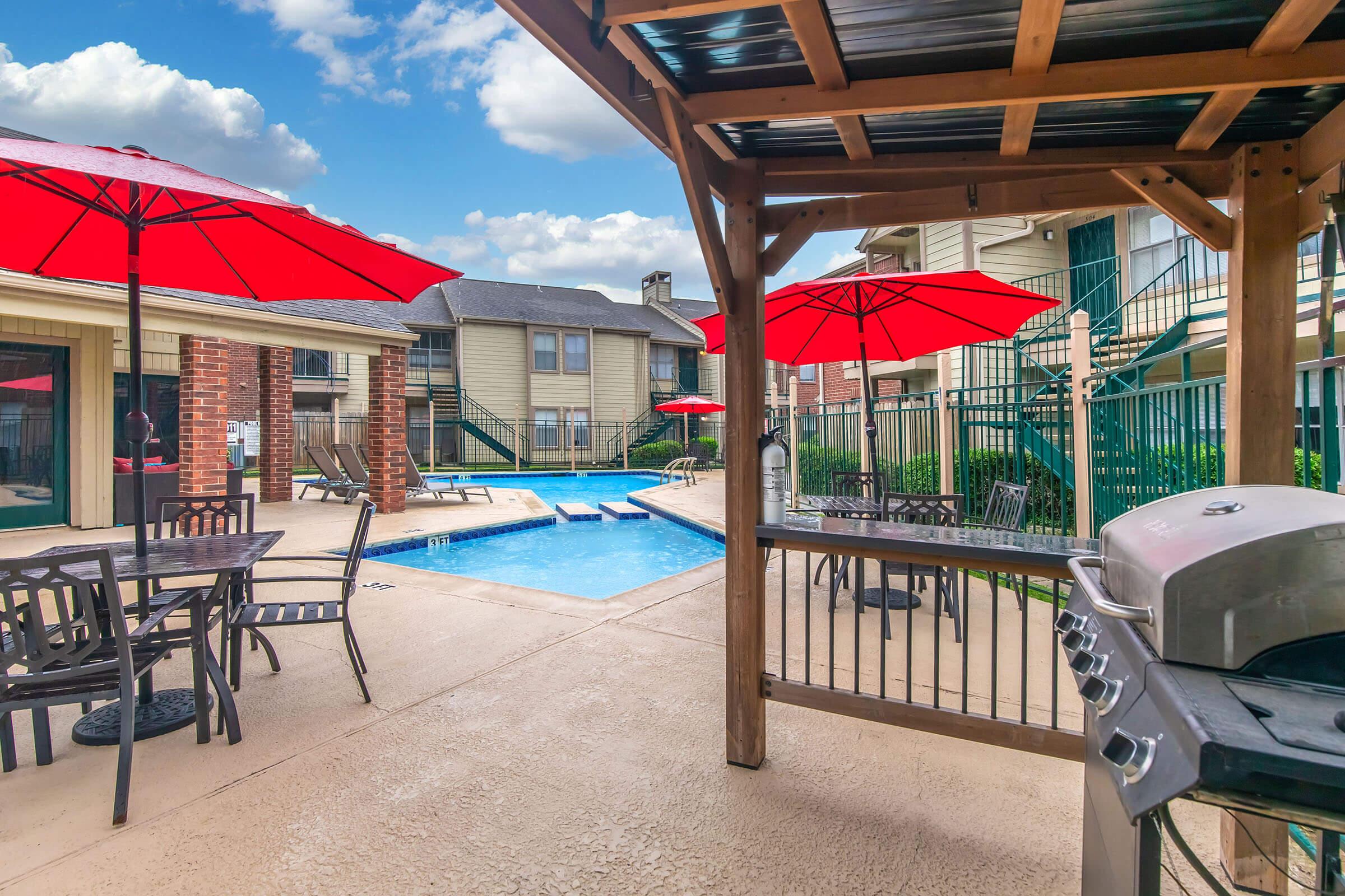 A pool area with lounge chairs and tables shaded by red umbrellas. A wooden gazebo structure features a grill, overlooking a clear blue pool surrounded by apartment buildings and green landscaping under a partly cloudy sky.