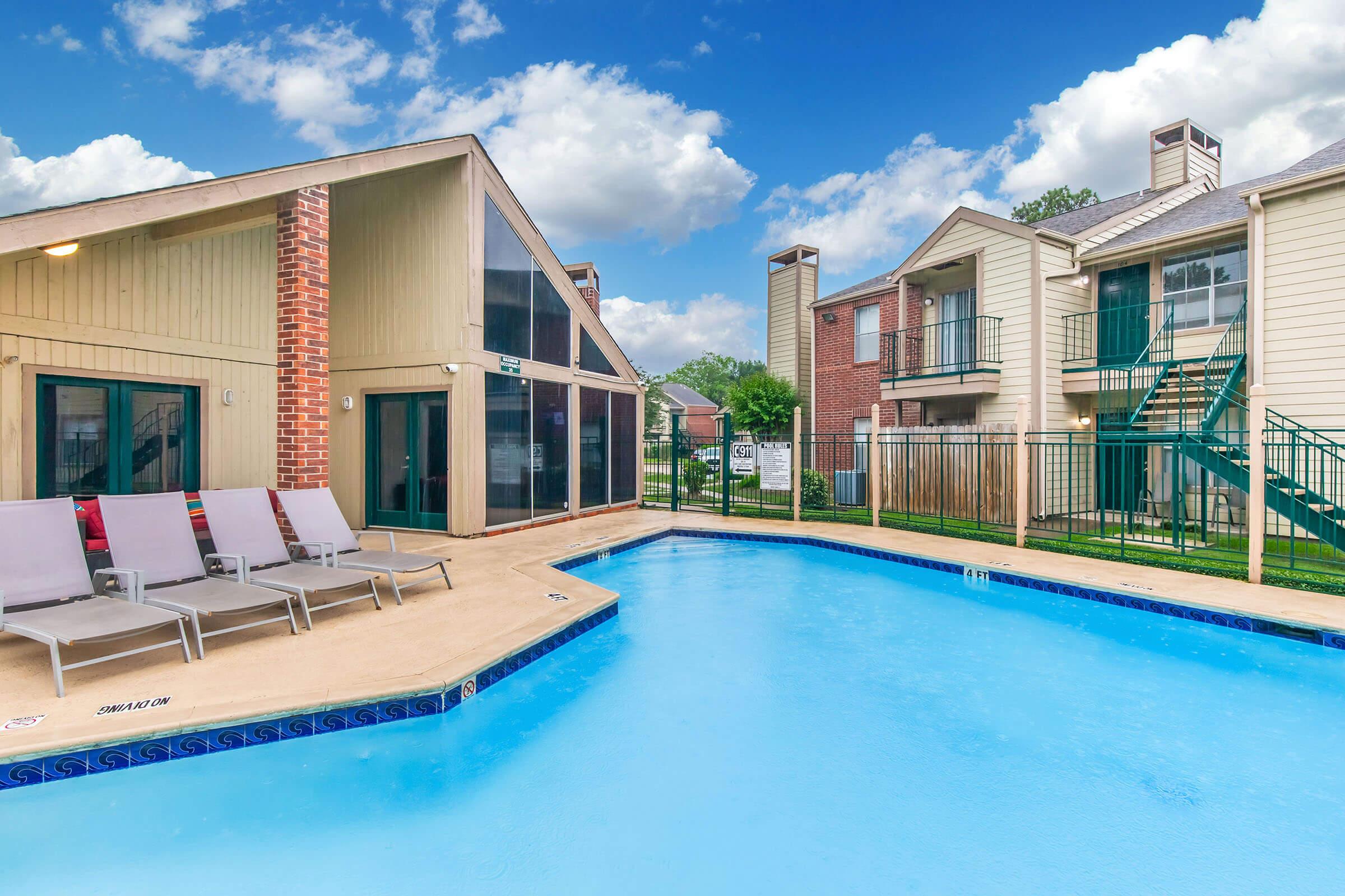 A view of a swimming pool surrounded by lounge chairs, with two multi-story residential buildings in the background. The pool area features a clean, blue surface, and the sky is partly cloudy, enhancing the inviting atmosphere of the outdoor space.