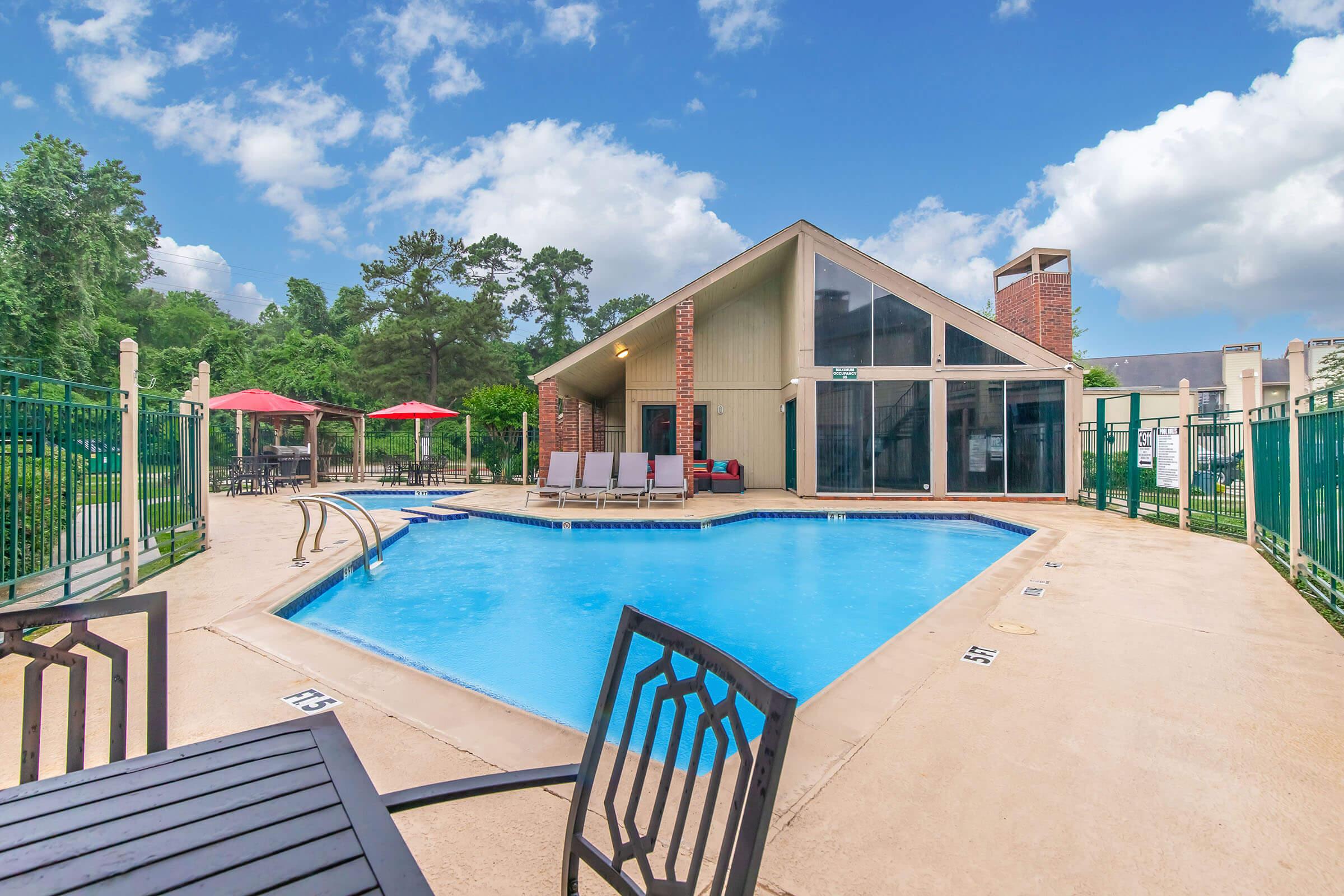 A tranquil outdoor swimming pool surrounded by a fenced area, featuring lounge chairs and red umbrellas. A modern building with large windows is adjacent to the pool, set against a backdrop of greenery and a partly cloudy blue sky.