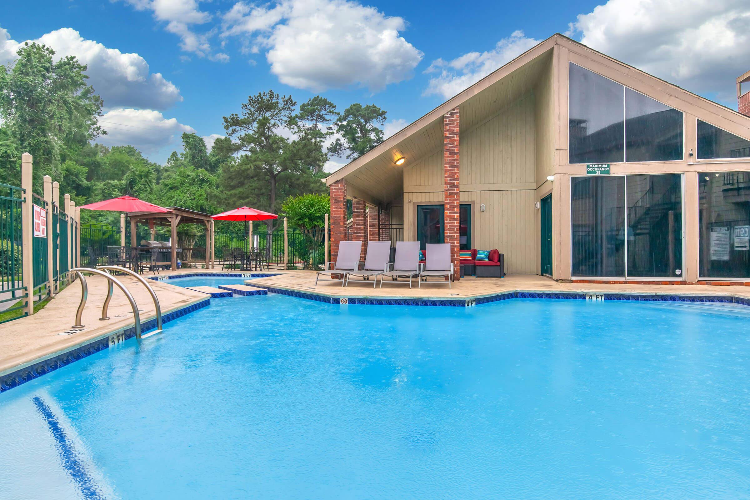 A serene outdoor pool scene featuring a crystal-clear swimming pool with a splash area, surrounded by a wooden deck. Lounge chairs sit beside the pool, and there are shaded seating areas with red umbrellas in the background. In the distance, a modern building with large windows complements the tranquil setting under a partly cloudy sky.