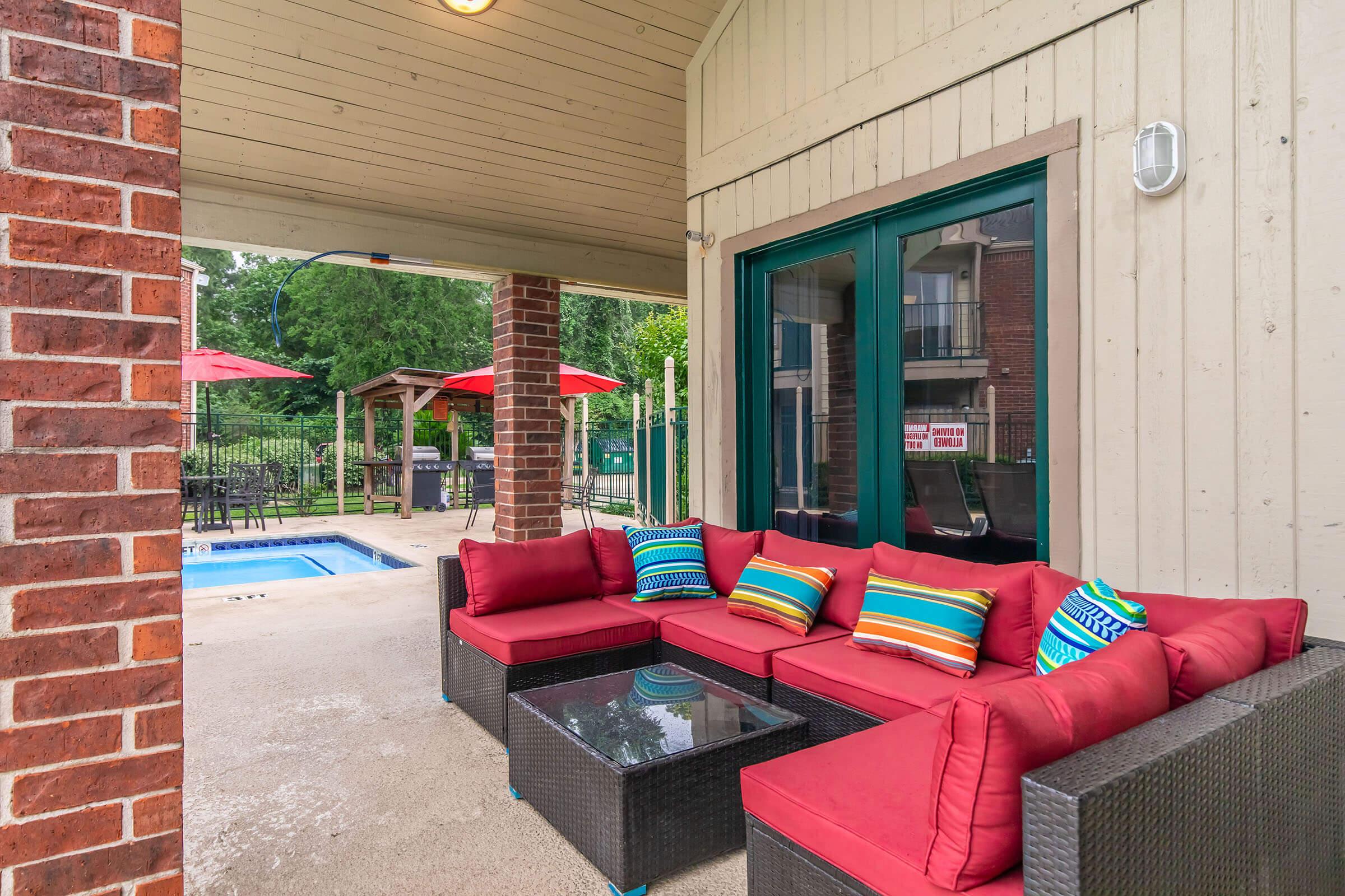 A patio area featuring a red sectional couch with colorful pillows, a glass coffee table, and a view of a pool in the background. There are also umbrellas providing shade and green landscaping, creating a relaxing outdoor space.