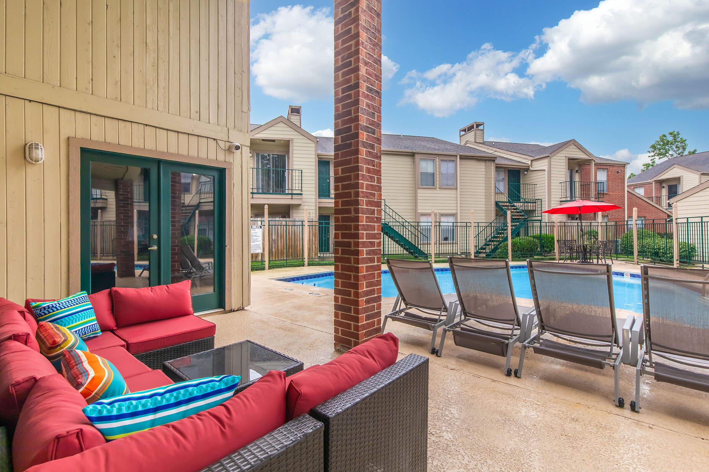 Outdoor pool area featuring a swimming pool surrounded by lounge chairs and a bright red umbrella. A cozy seating area with red cushions and colorful pillows is visible, along with a building facade and green landscaping in the background. Cloudy blue sky above.
