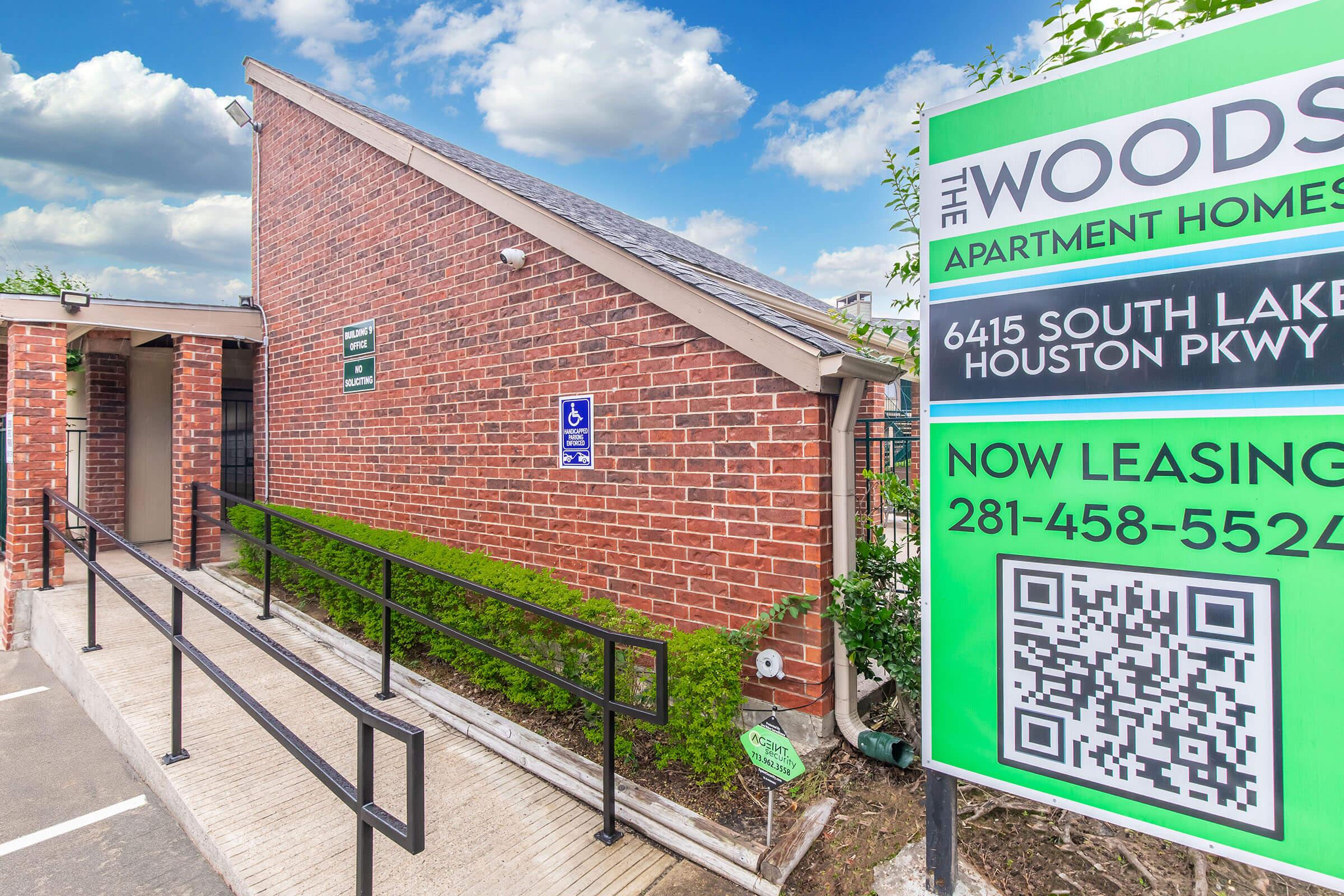 Exterior view of The Woods Apartment Homes, featuring a brick building with a sloped roof, a wheelchair-accessible ramp, and a green sign with leasing information. The sign includes a QR code and contact number for inquiries. Clear blue sky with some clouds in the background.
