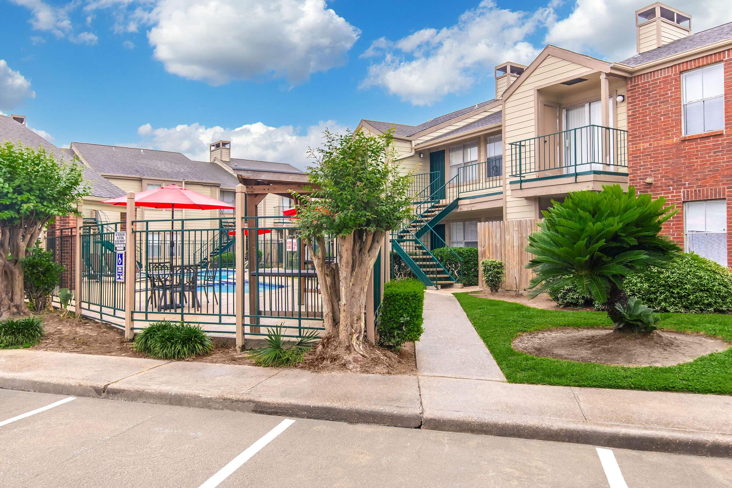 A well-maintained apartment complex featuring two-story buildings with a green lawn, shrubs, and trees. In the foreground, there is a fenced pool area surrounded by lounge chairs and red umbrellas. The sky is partly cloudy, adding a bright backdrop to the scene.