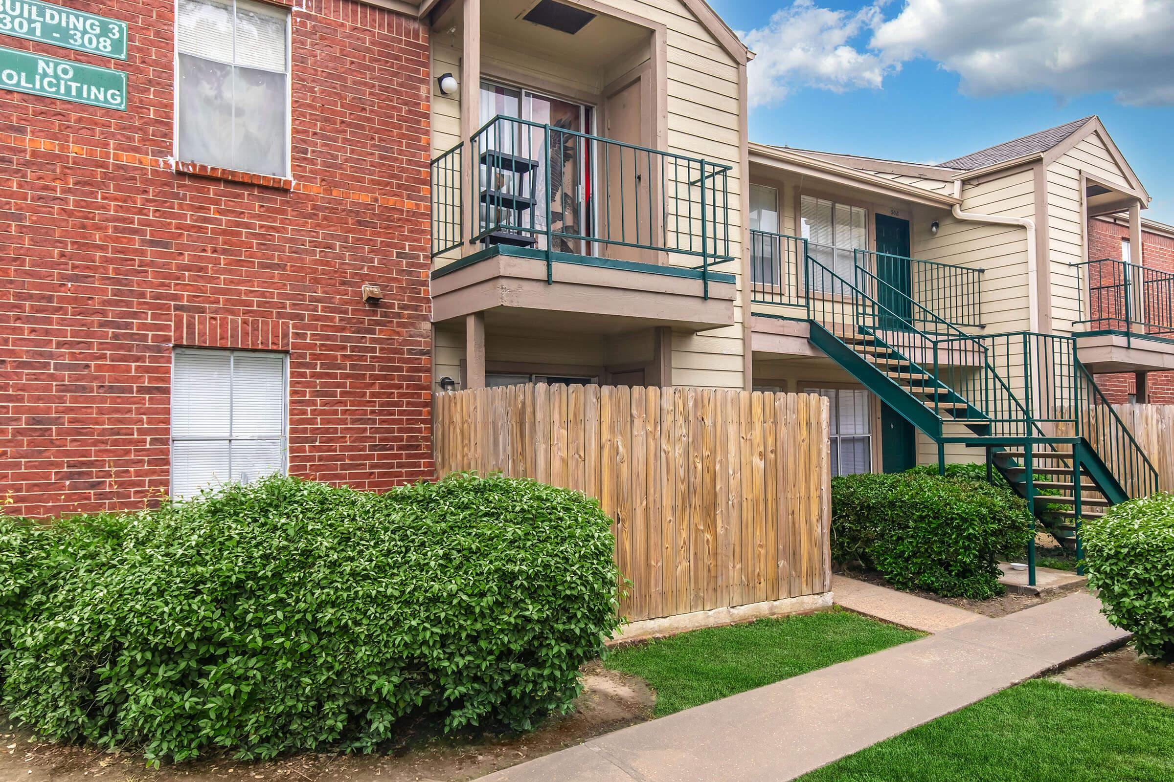 A two-story apartment building with red brick and beige siding. There are green railings on the balconies, and a wooden fence separates the building from lush green bushes. The sidewalk leads up to the entrance, and a sign on the building reads "Building 3." The sky is partly cloudy.