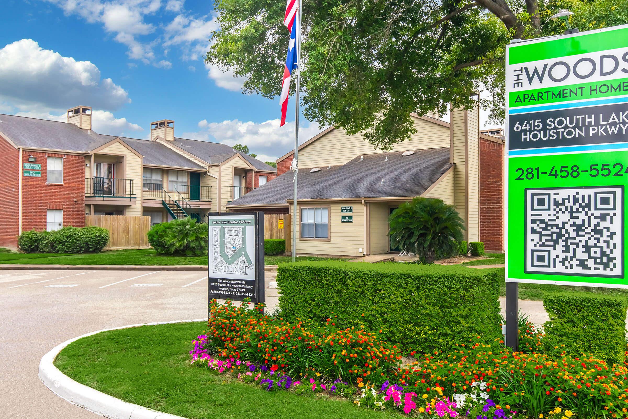 Image of the Woods Apartment Homes entrance, featuring a sign with contact information and a QR code. The surroundings include landscaped greenery, flower beds, and multiple apartment buildings in the background. A Texas flag is also visible. The scene is set on a clear day with a blue sky.