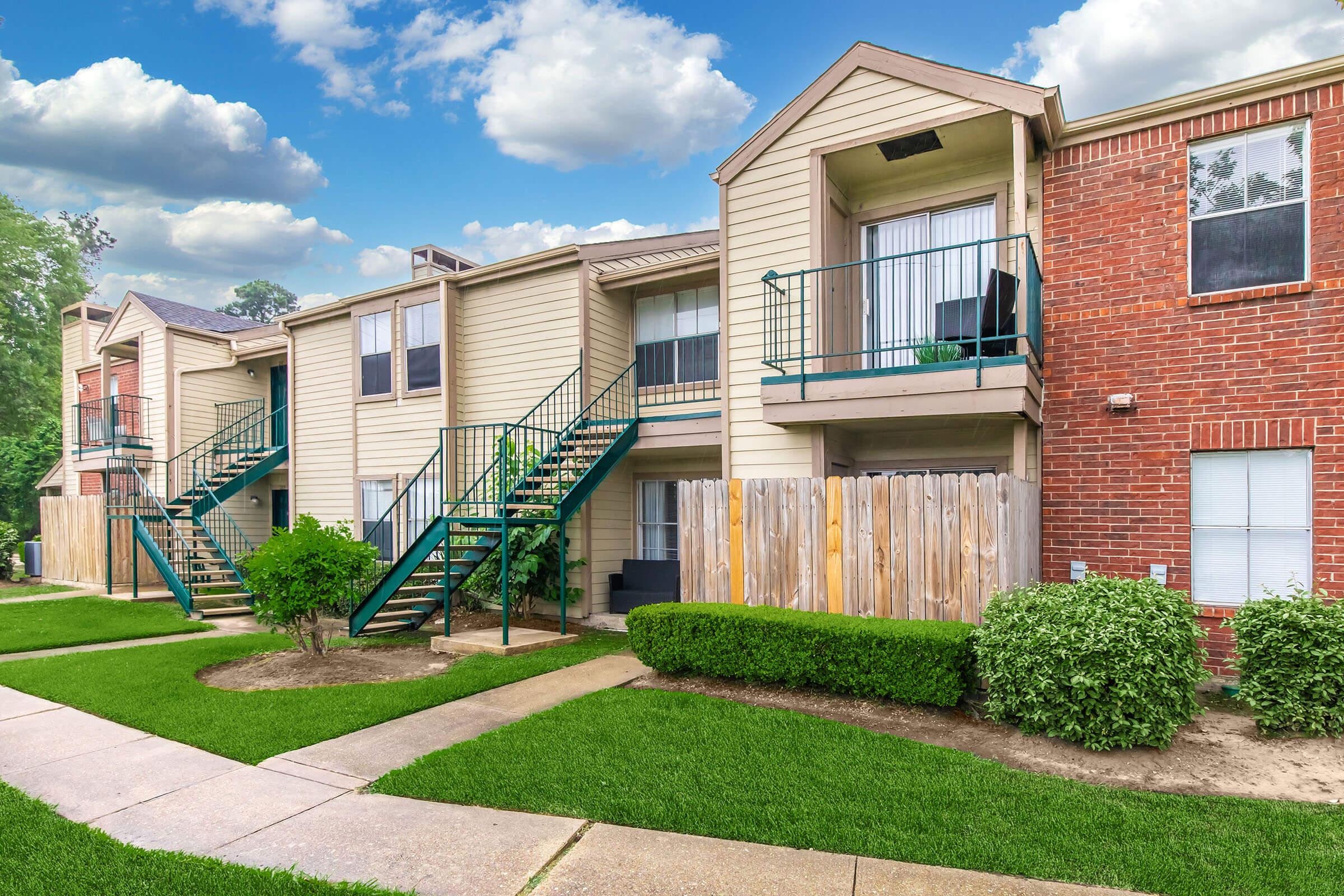 A well-maintained apartment complex featuring two-story buildings with a mix of brick and siding exteriors. Green staircases lead to the upper units, and small balconies are visible. The landscaped grounds include manicured lawns and tidy hedges, with a sidewalk running along the front of the buildings. Cloudy blue sky above.