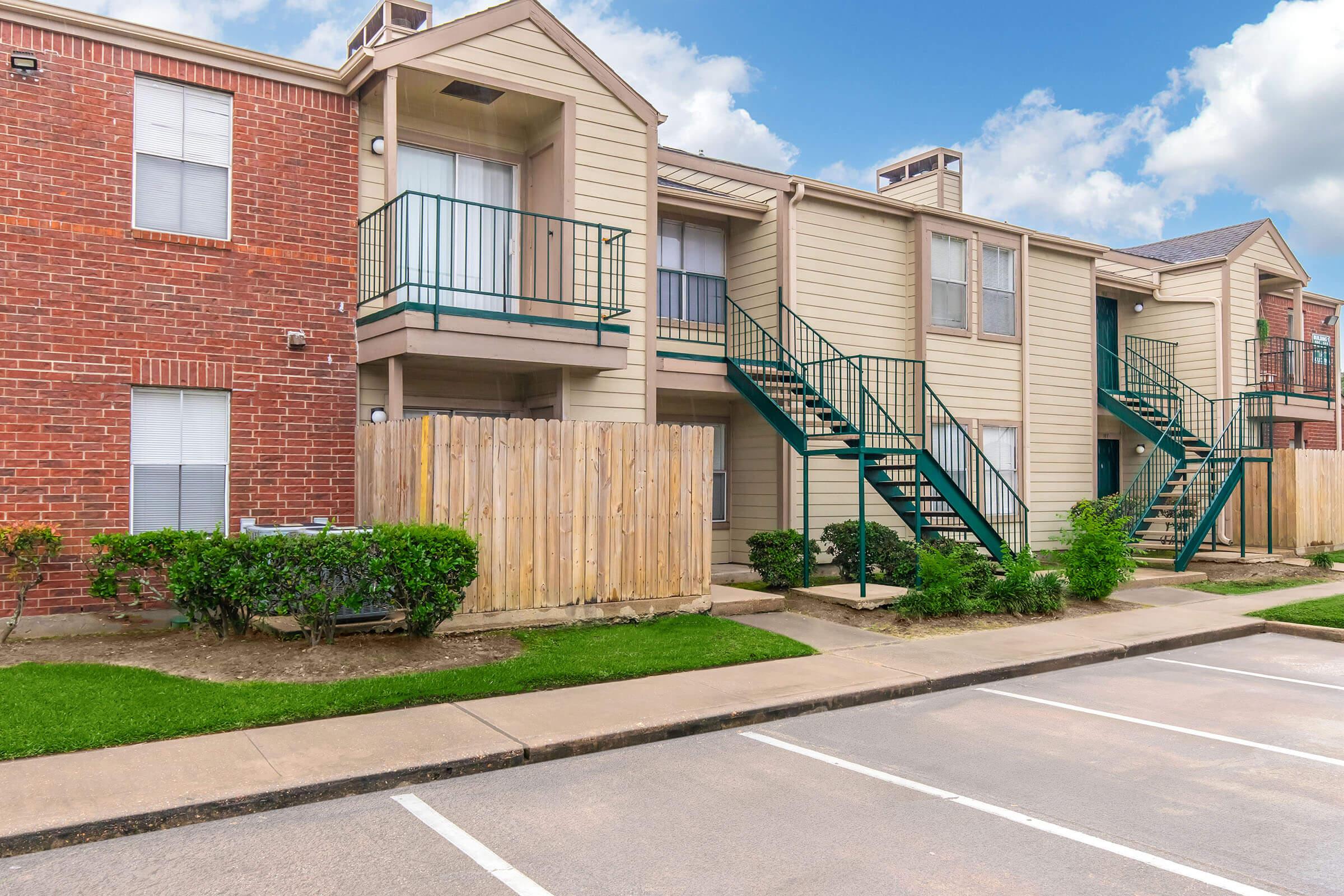 Two-story apartment building with a mix of brick and beige siding. Green metal staircases lead to the upper units. A wooden fence surrounds a small yard with landscaping. The parking lot is empty with designated parking spaces visible. The sky is partly cloudy, giving a bright atmosphere to the scene.