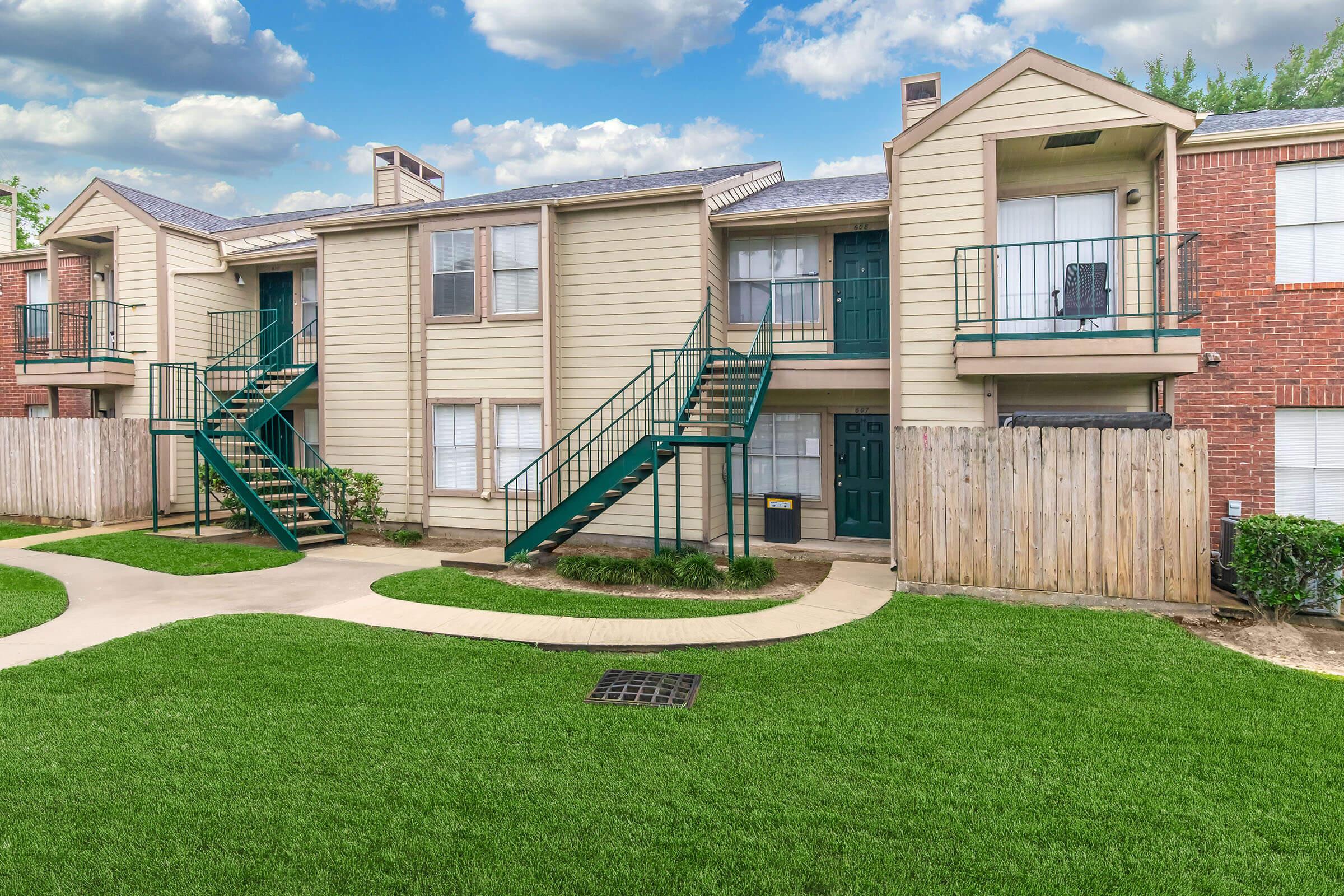 A two-story apartment building with light-colored siding, green staircases, and a fenced yard. The lawn is lush and well-maintained, with a winding pathway leading to the entrance. Soft clouds can be seen in the blue sky above.