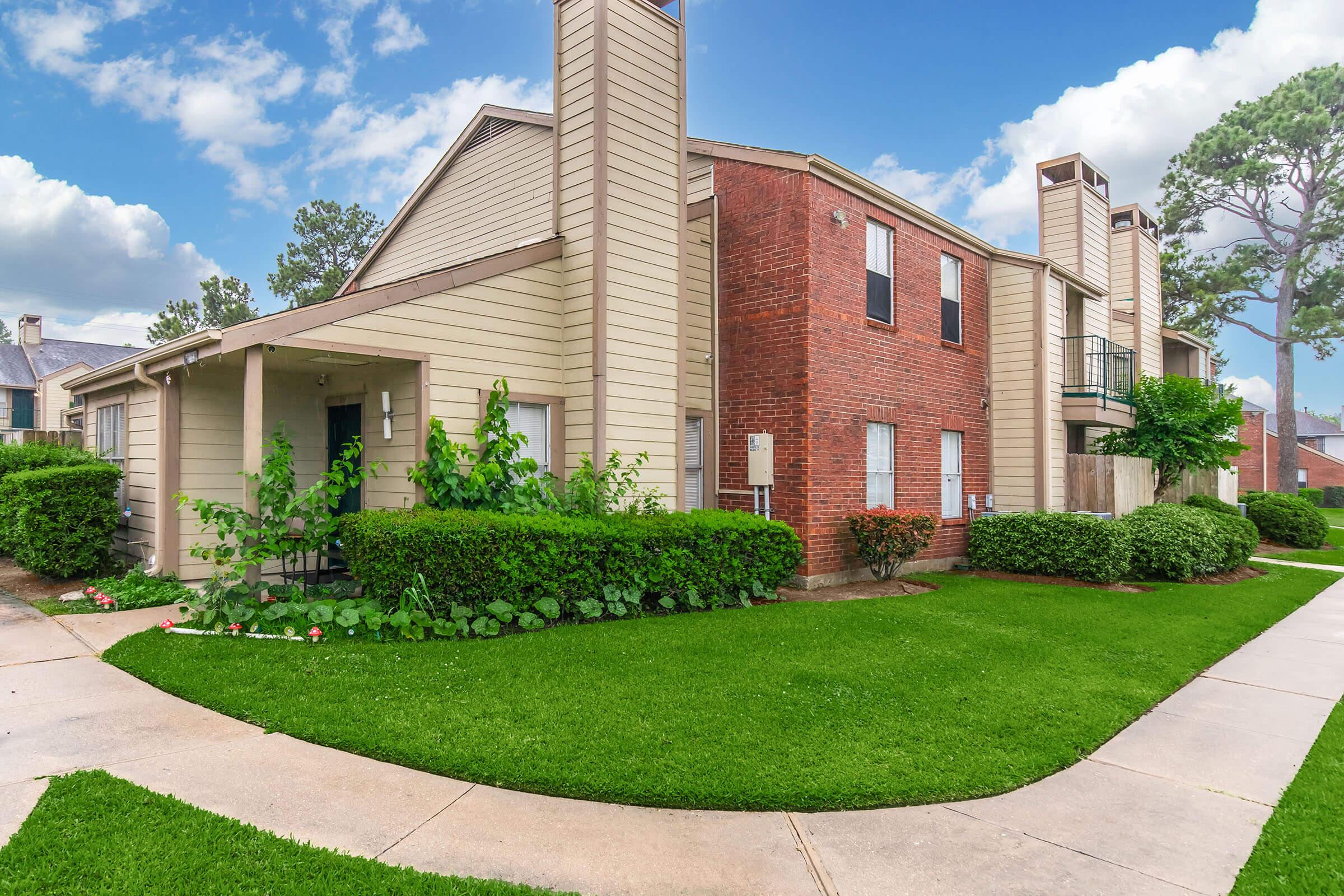 A well-maintained two-story apartment building featuring a mix of light-colored siding and brick. It has green shrubs and neatly trimmed grass surrounding the building, with a curved concrete walkway leading up to the entrance. Bright blue skies with a few clouds are visible in the background.