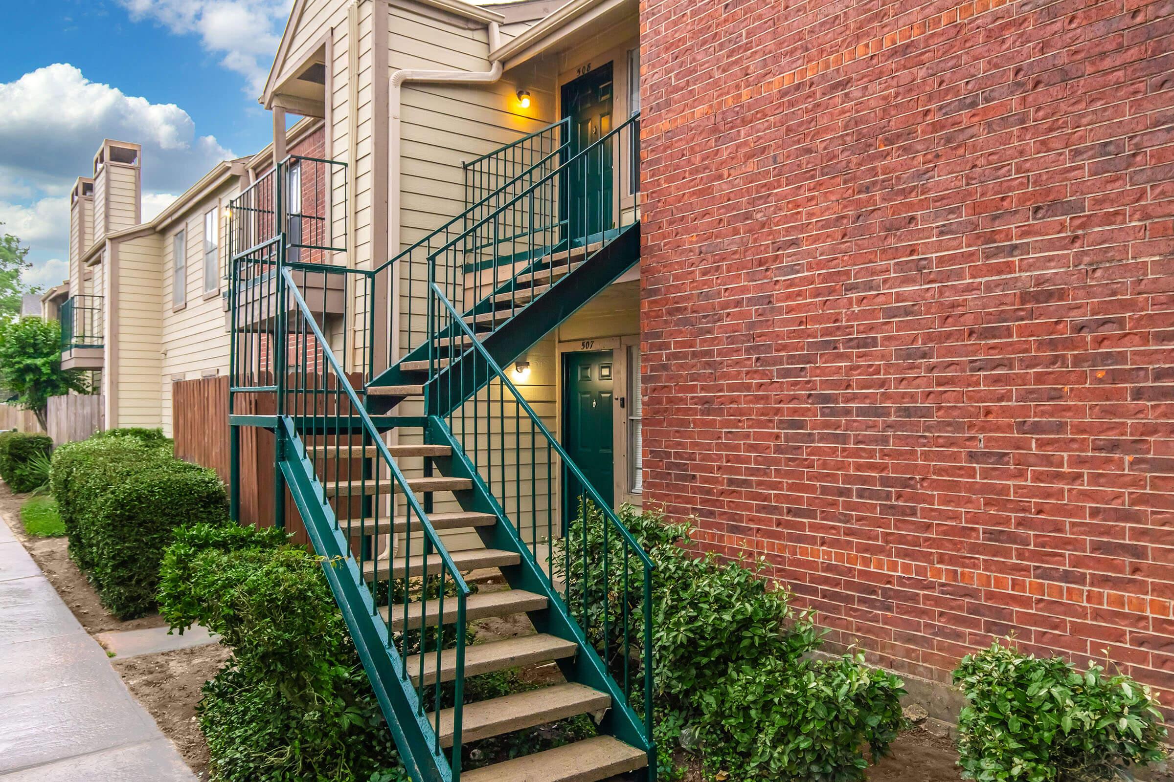 Exterior view of an apartment building featuring a set of green metal stairs leading to upper units. The building has a combination of light beige siding and red brick. Lush green bushes line the walkway, and the sky above is partly cloudy.