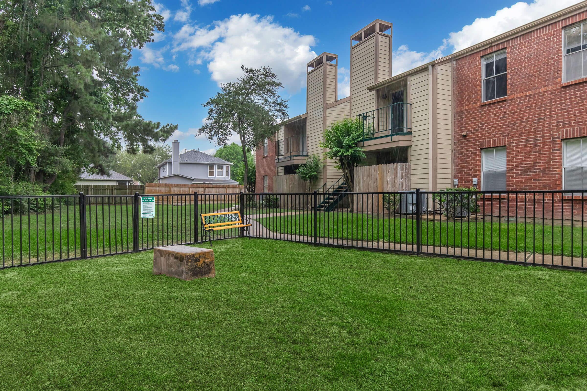 A well-maintained outdoor area featuring a grassy lawn enclosed by a black fence. In the center, there's a wooden bench and a tree stump. Two multi-story apartment buildings are visible in the background, with several windows and a small landscaped area. The sky is partly cloudy, suggesting a pleasant day.