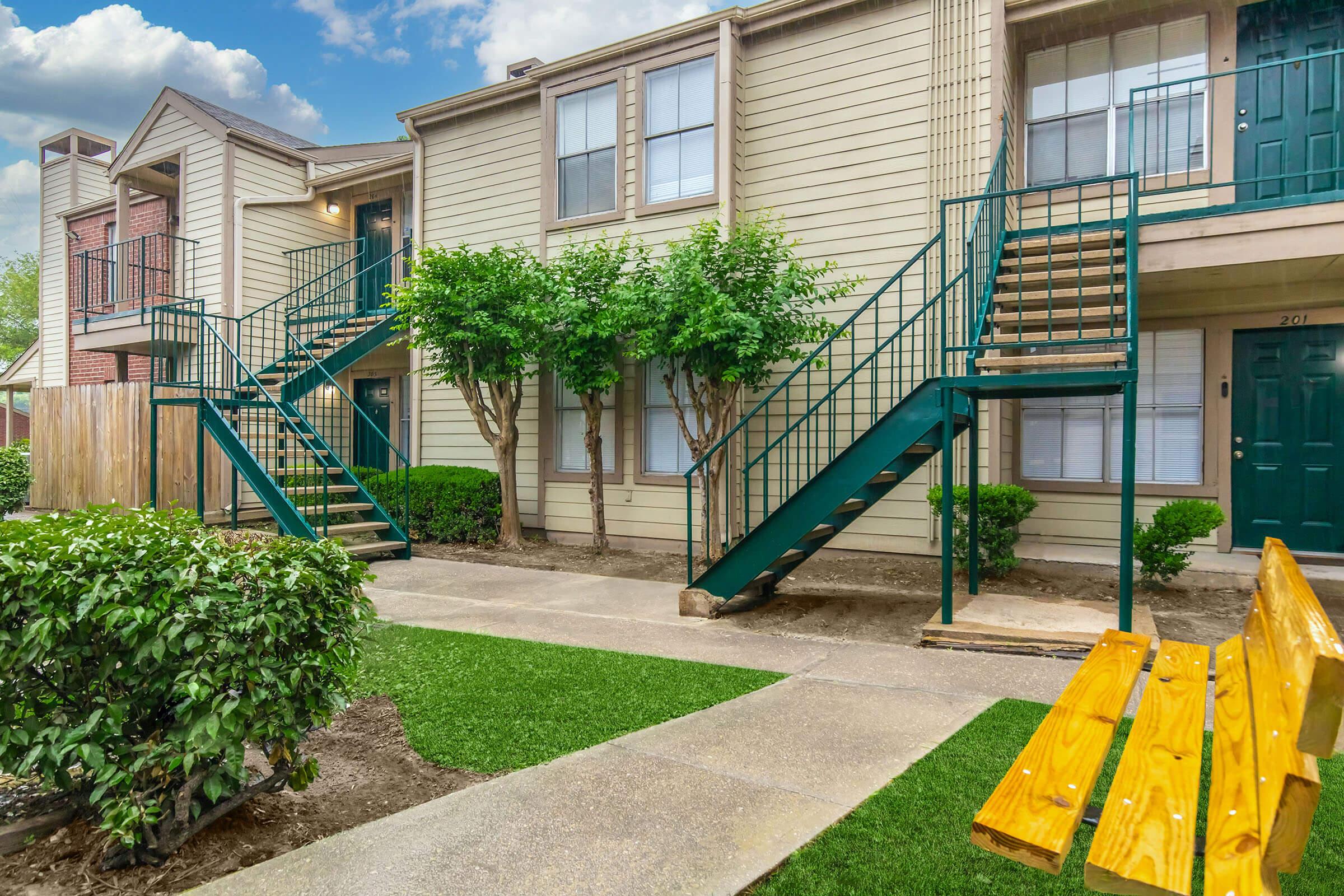 A well-maintained apartment complex featuring two-story buildings with green staircases. Lush bushes and neatly trimmed grass line the walkway, which leads to the entrance of the apartments. A wooden bench is positioned nearby for seating. The sky is partly cloudy, adding to the pleasant atmosphere.